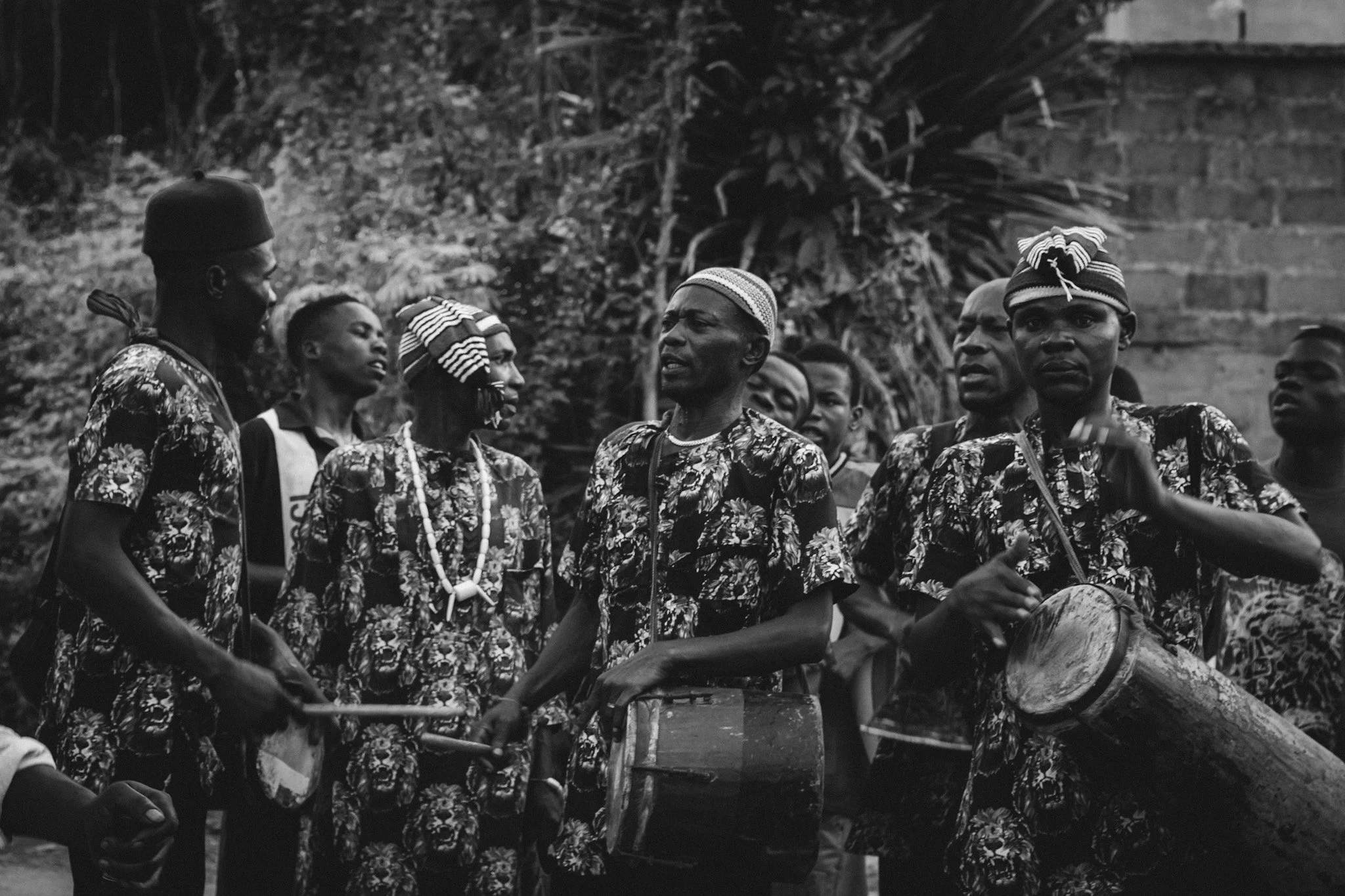 A group of men dressed in traditional African clothing, some with hats and beaded necklaces, gather outdoors, standing near palm trees and a brick wall, engaging in conversation.