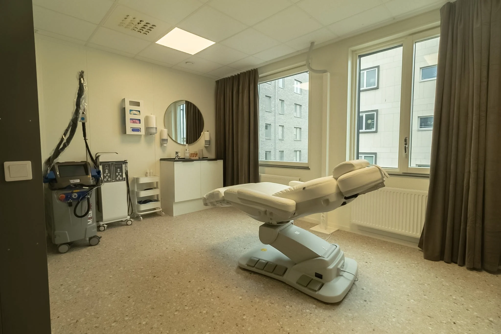 A hospital room with a medical bed, medical equipment, large windows with curtains, and city buildings outside.