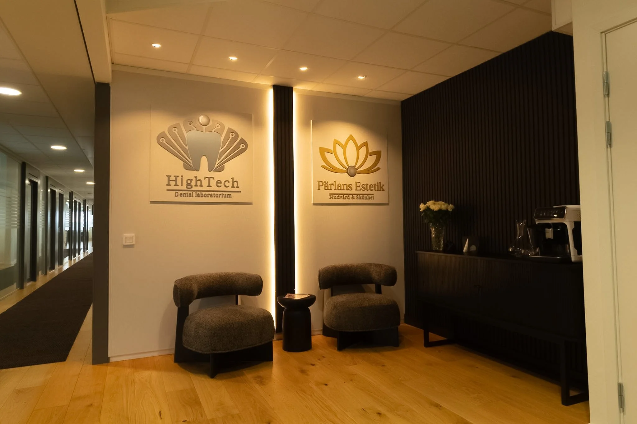 Interior of an office or waiting area with two signs on the wall, one for 'HighTech Dental Laboratorium' and the other for 'Pärlans Estetik Hårvård & Skönhet,' two chairs, a small table, a vase with flowers, and a coffee machine.