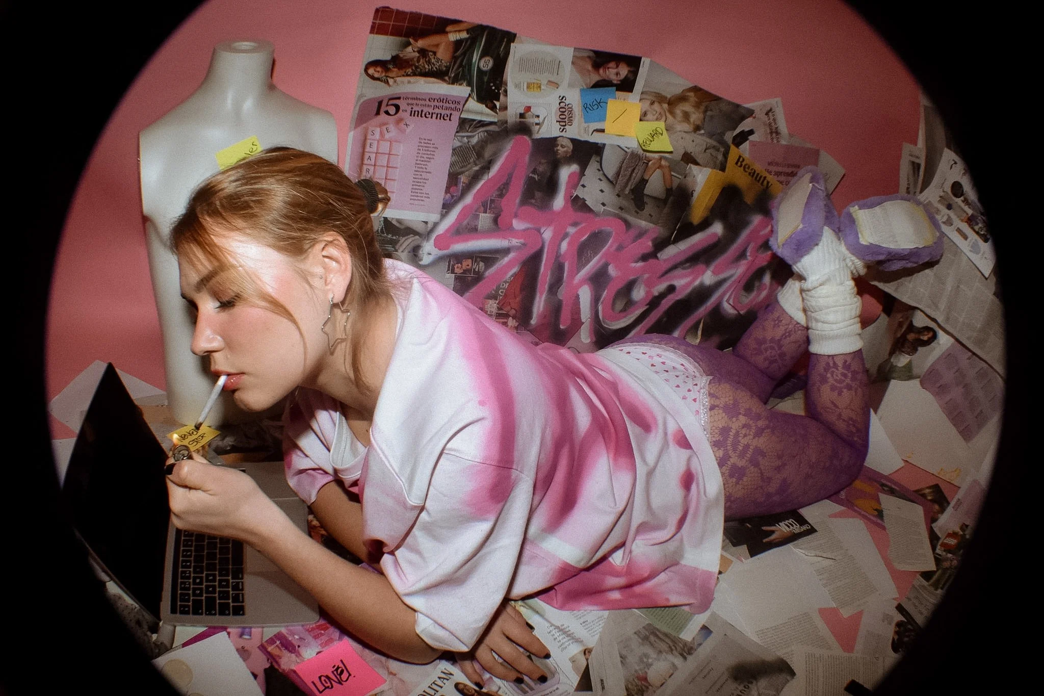 A woman with necklace earrings, lying on her stomach on a pink background, surrounded by magazines, newspapers, and sticky notes. She is holding a pen and appears to be writing or drawing.