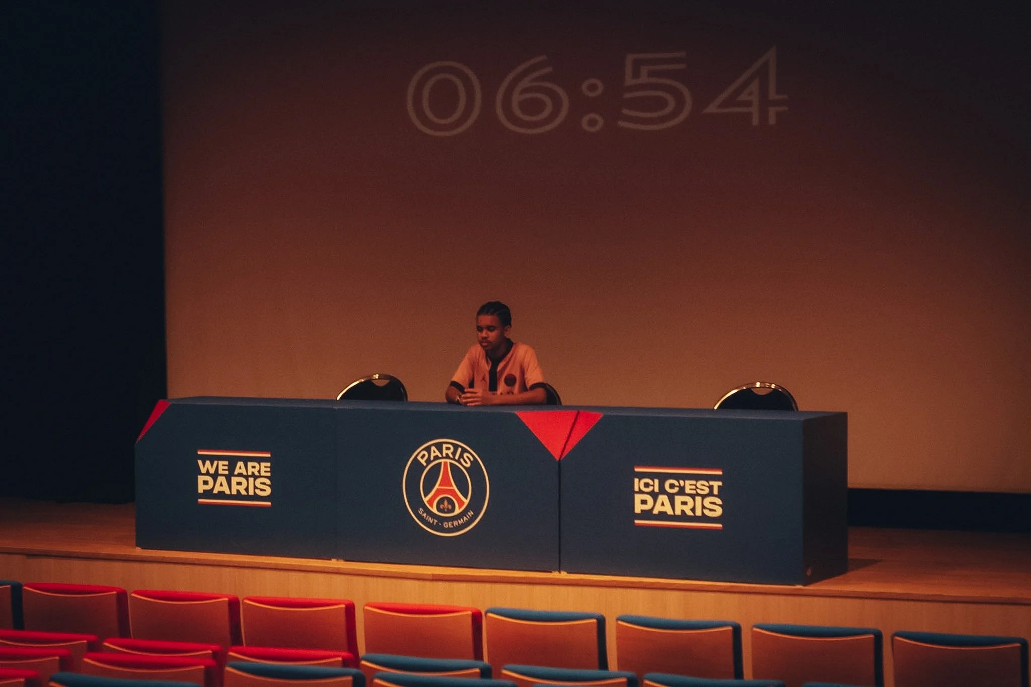 A young person sitting behind a table with Paris Saint-Germain logos and slogans, in a dimly lit auditorium with red and blue chairs in the foreground.