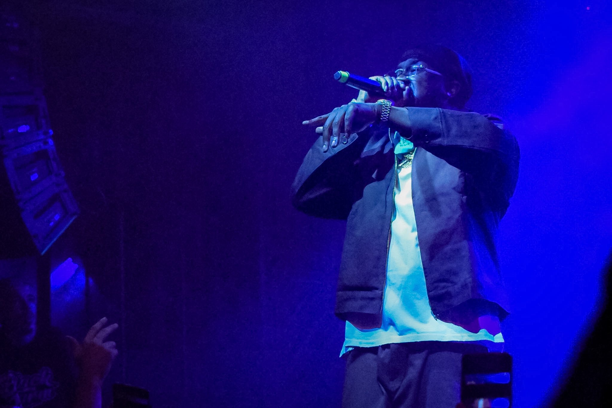 A person performing on stage with a microphone, illuminated by blue stage lighting, wearing glasses, a dark jacket, and a watch.