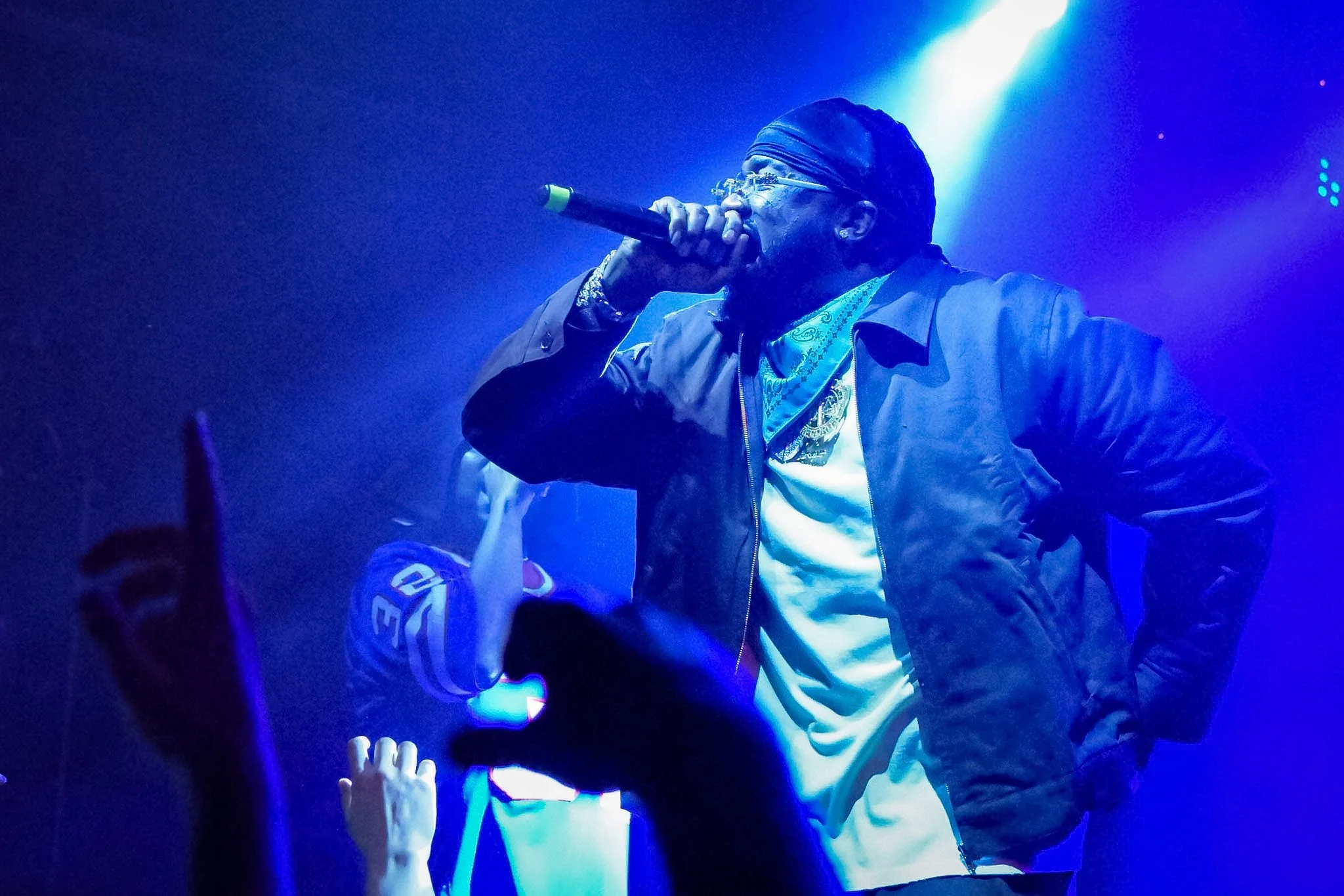 Performer singing into a microphone on stage, wearing glasses, a bandana, a jacket, and a patterned shirt, in a concert setting with blue and purple lighting, and audience members visible at the bottom.