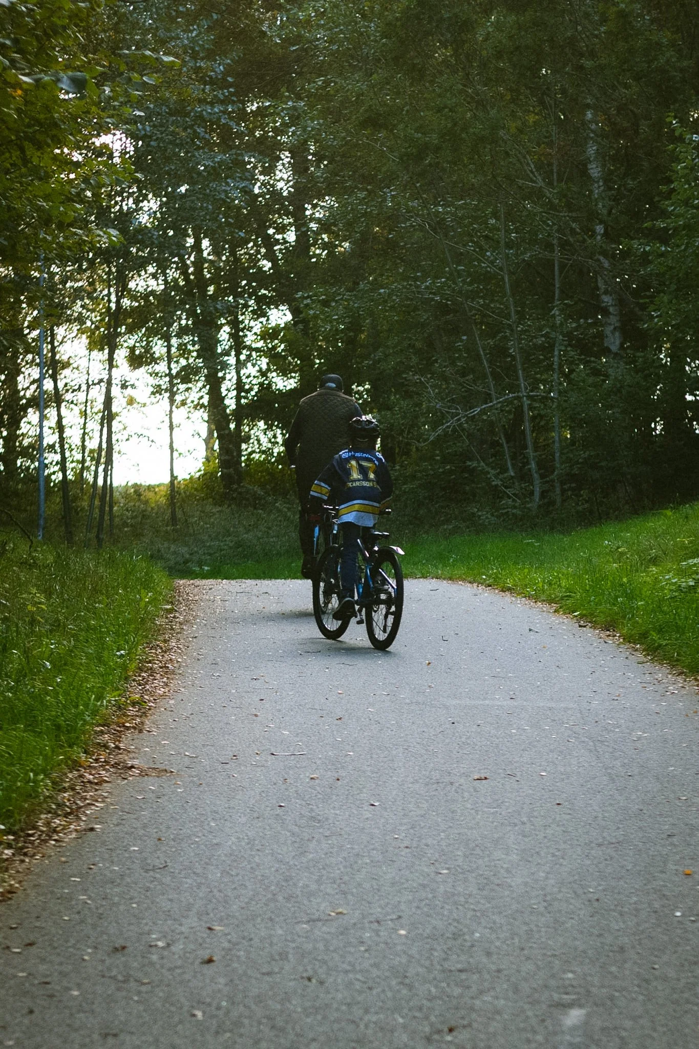 Two children riding bikes on a paved path through a wooded area, with trees on either side and light filtering through the leaves.