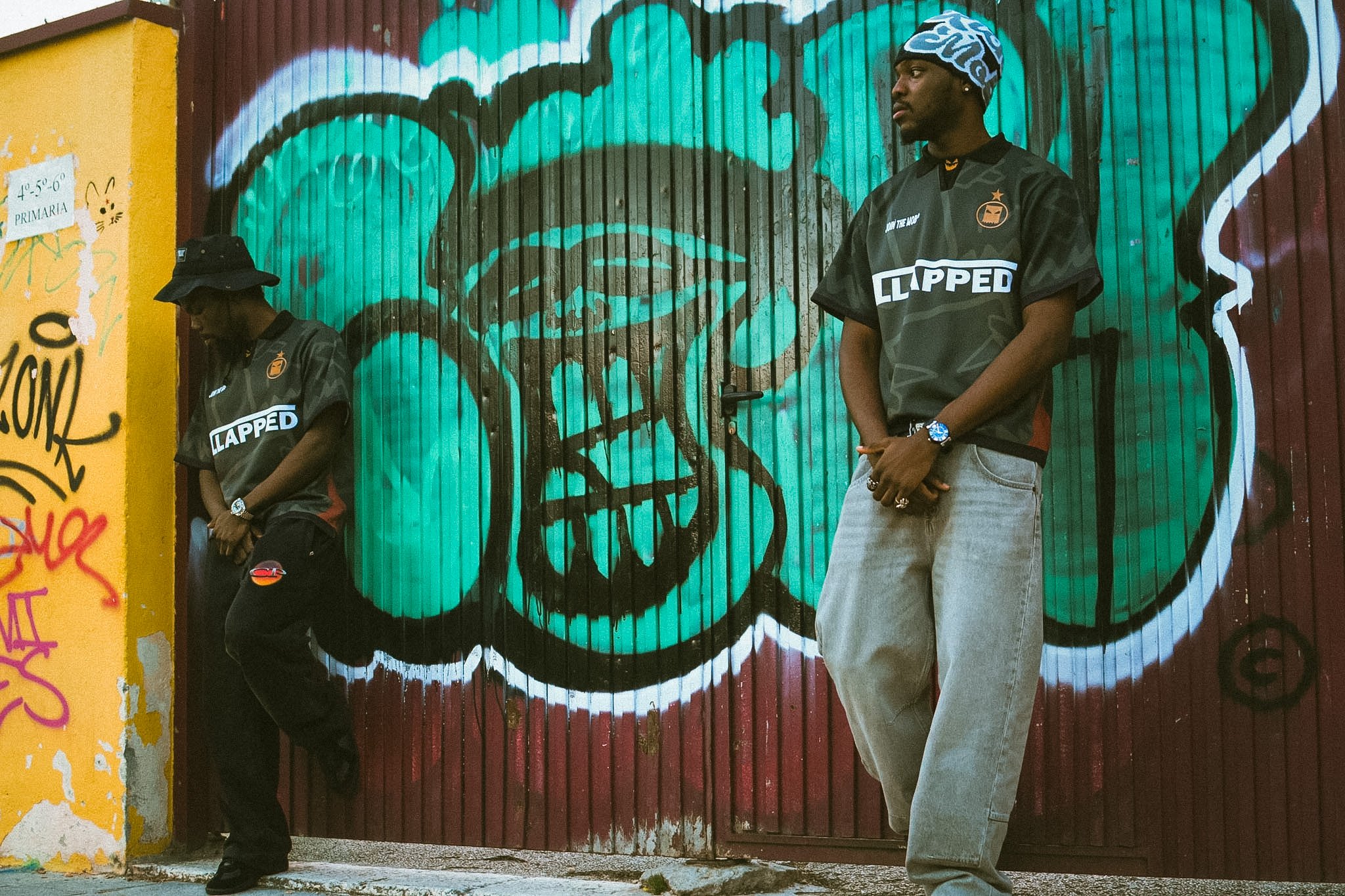 Two men stand in front of a graffiti-covered metal door with teal and black spray paint art. One man is wearing a black jacket with patch details, a watch, and black pants with a bucket hat. The other man is wearing a black T-shirt with a logo, a pri