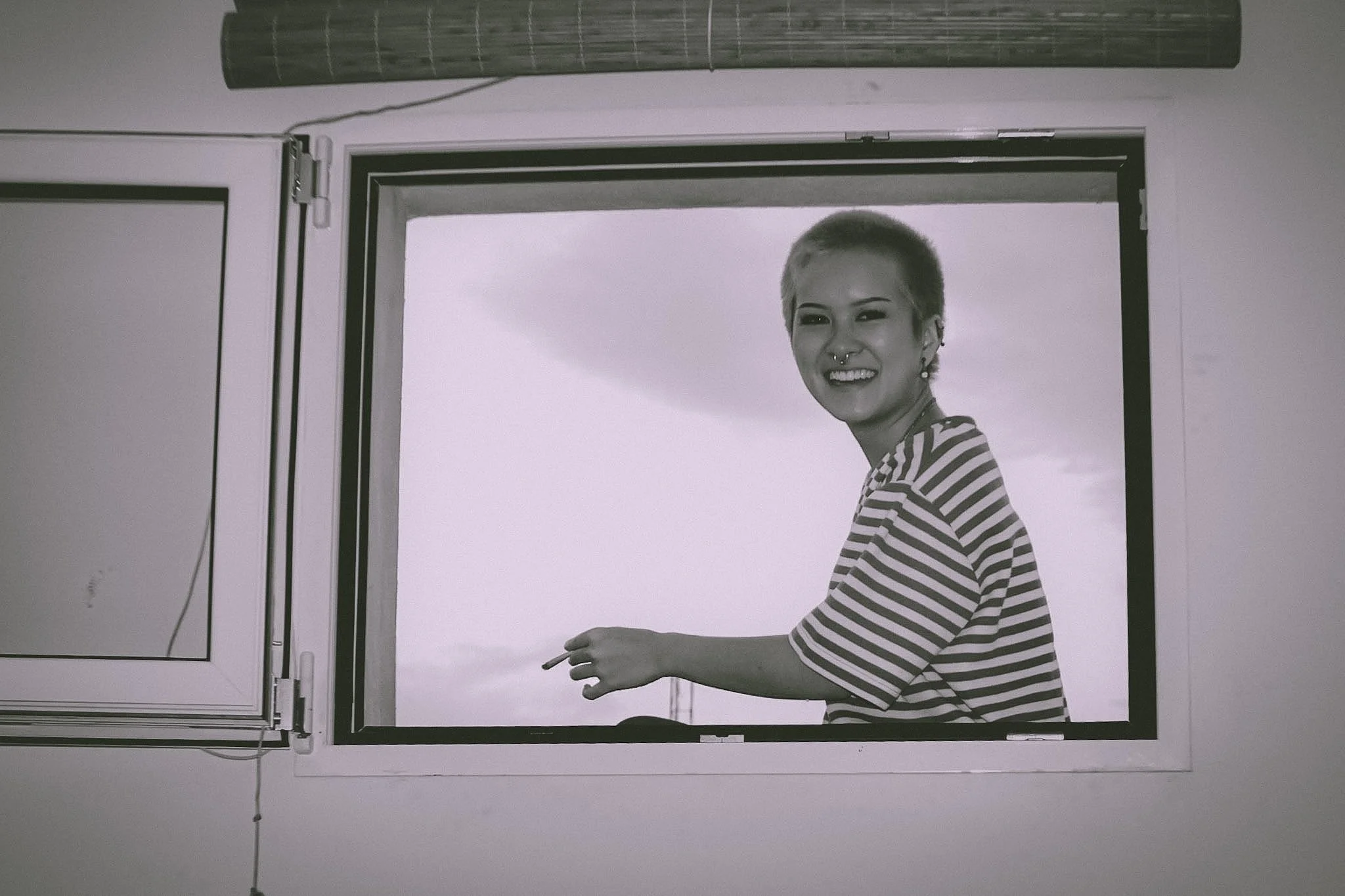 A person with short blonde hair and a septum piercing, wearing a black and white striped shirt, is standing in front of a window or glass door, smiling and looking towards the camera.
