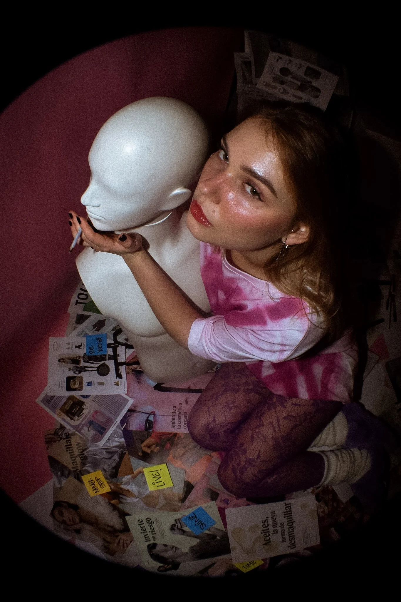 A woman with long hair, wearing a pink tie-dye shirt, purple lace tights, and striped socks, is holding a white mannequin head close to her face. The woman is smiling with shiny red lipstick, and there are various flyers, notes, and photos taped to a