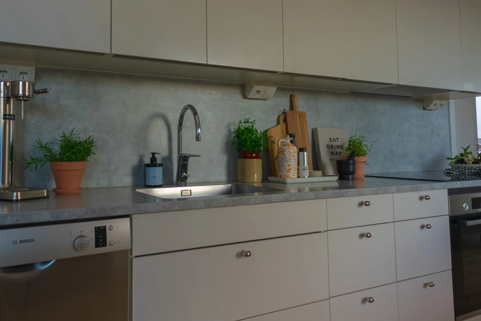 Kitchen countertop with potted herbs, cutting boards, soap dispenser, and a sign that says 'Eat Drink Nap'.