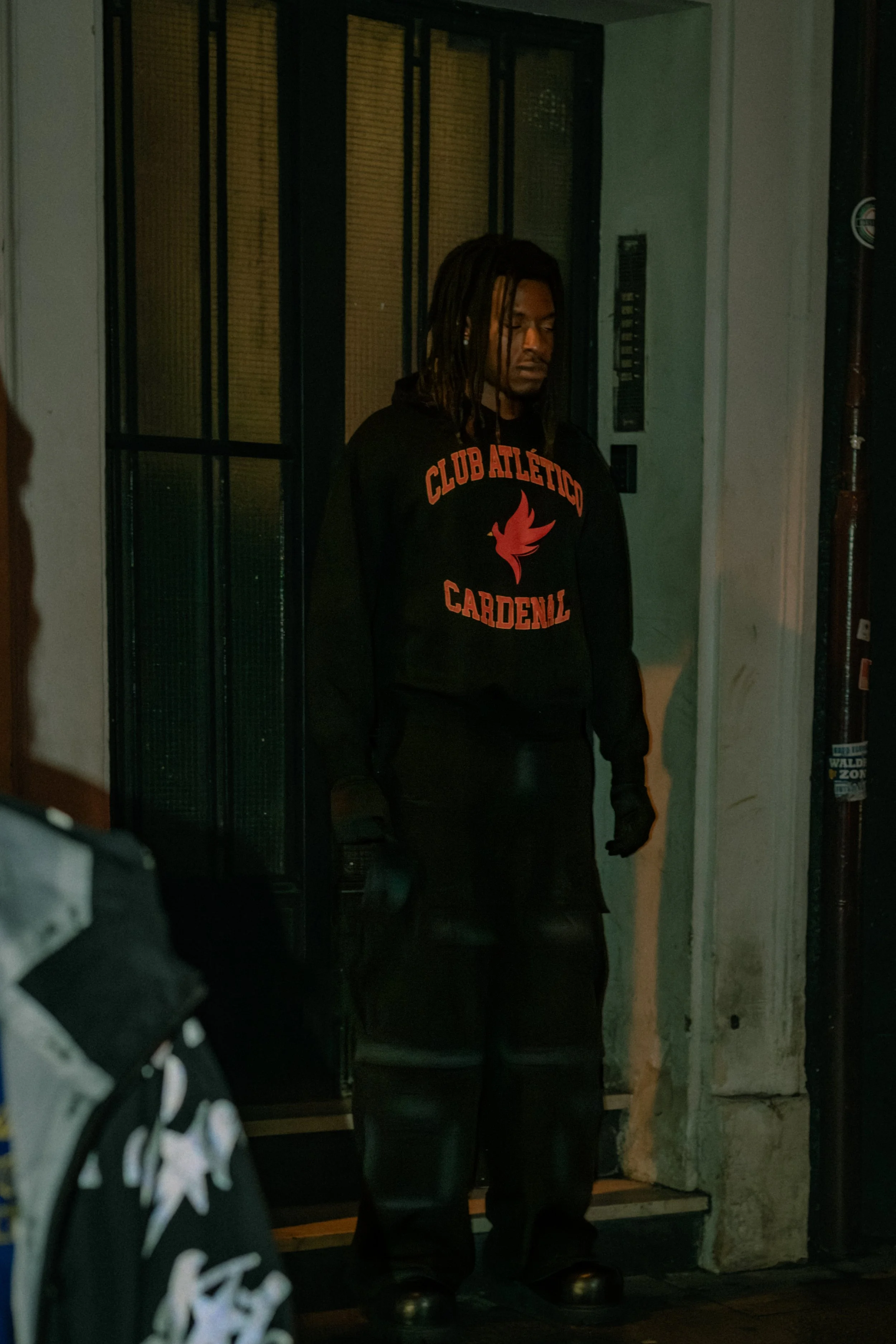 A person with dark skin and dreadlocks standing in front of an elevator, wearing all black with a hoodie that says 'Club Atlético Cardenal' and a red bird logo. The person has closed eyes and appears to be looking downward.