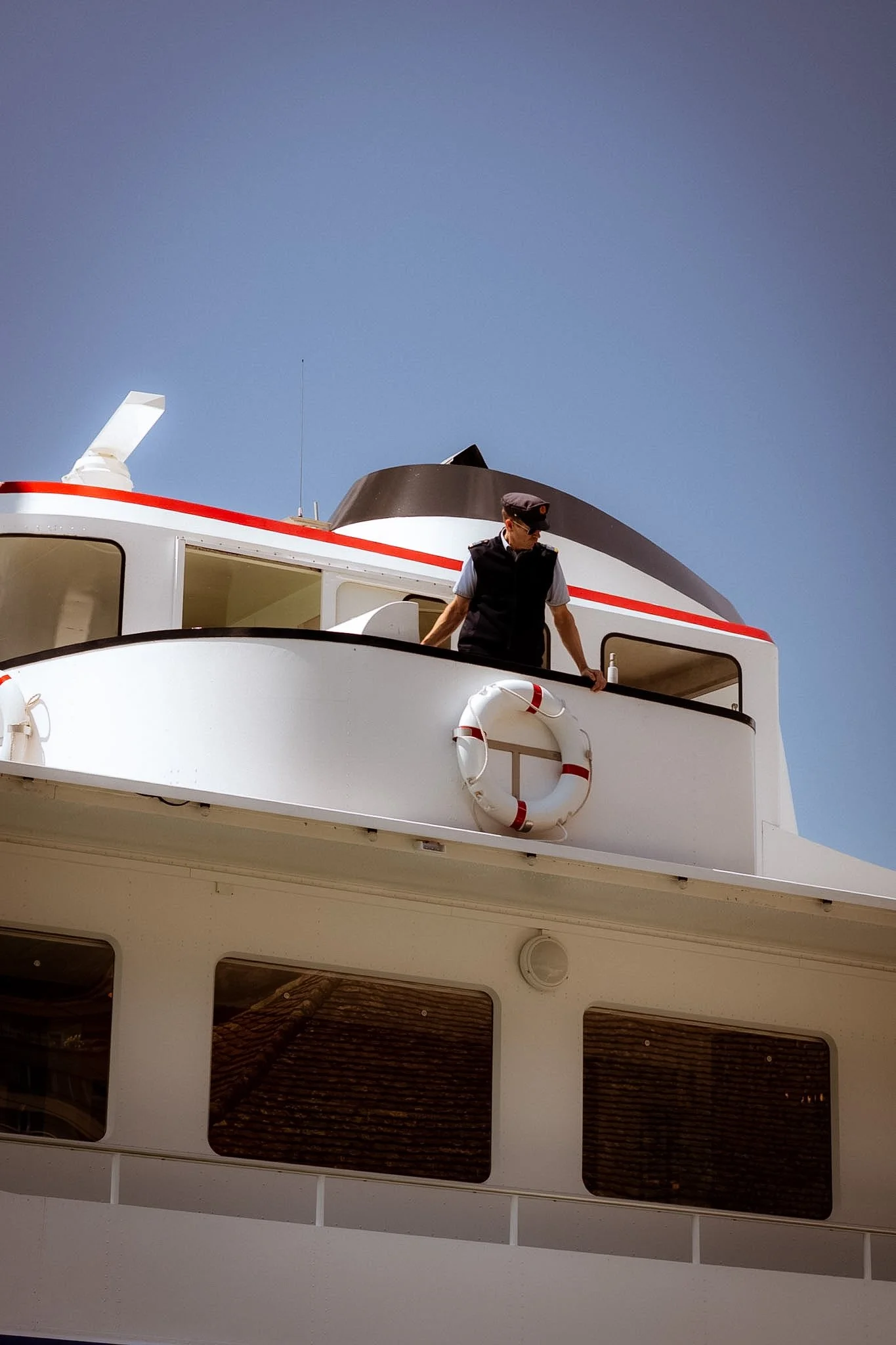 A man in a navy vest, white shirt, and navy cap stands on a balcony of a yacht, looking down. The yacht has white walls with large rectangular windows and a life preserver hanging on the side. The sky is clear blue.