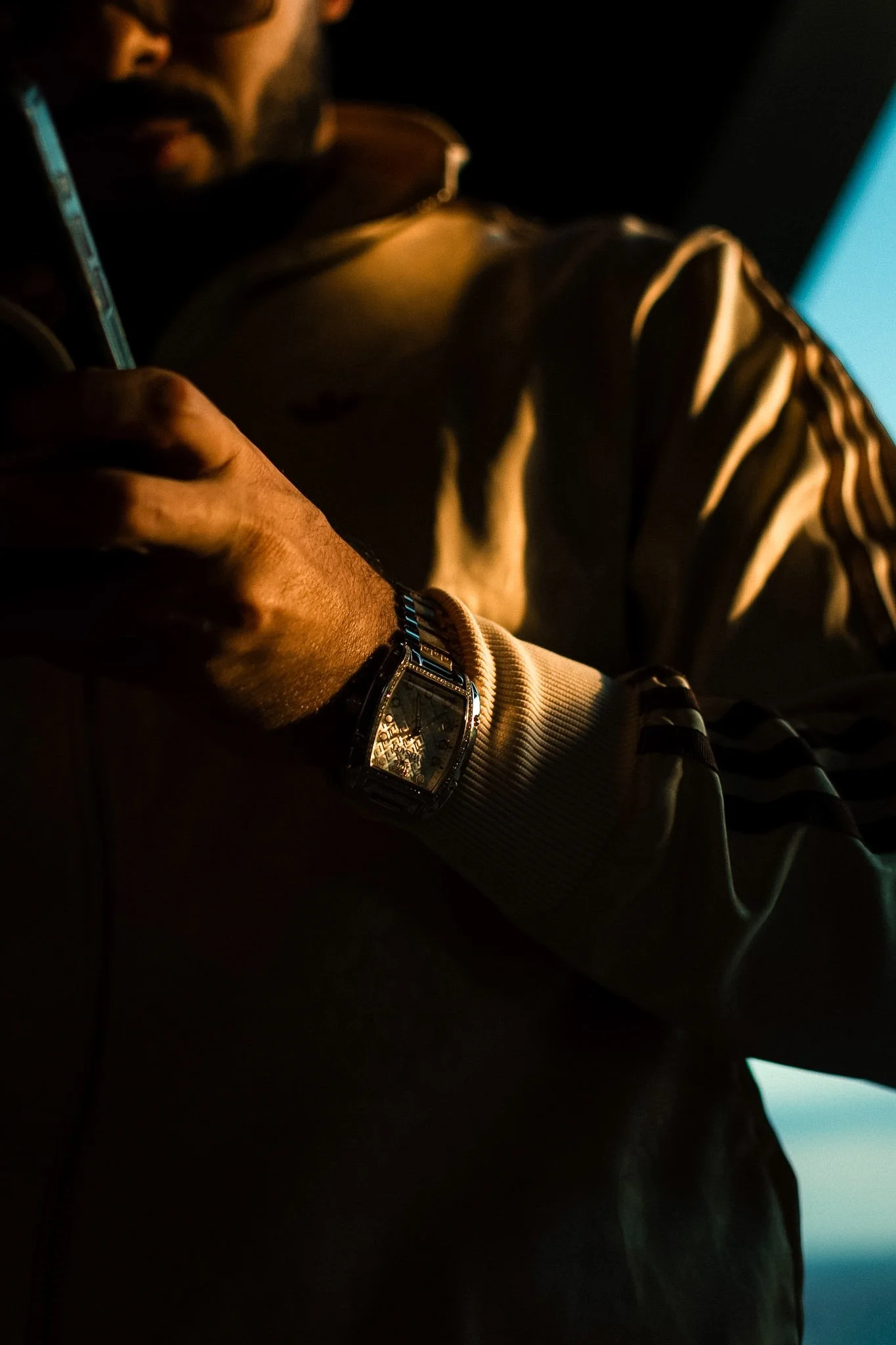 A person sitting indoors at night, wearing a watch and a striped long-sleeve shirt, looking at a screen emitting a blue light.
