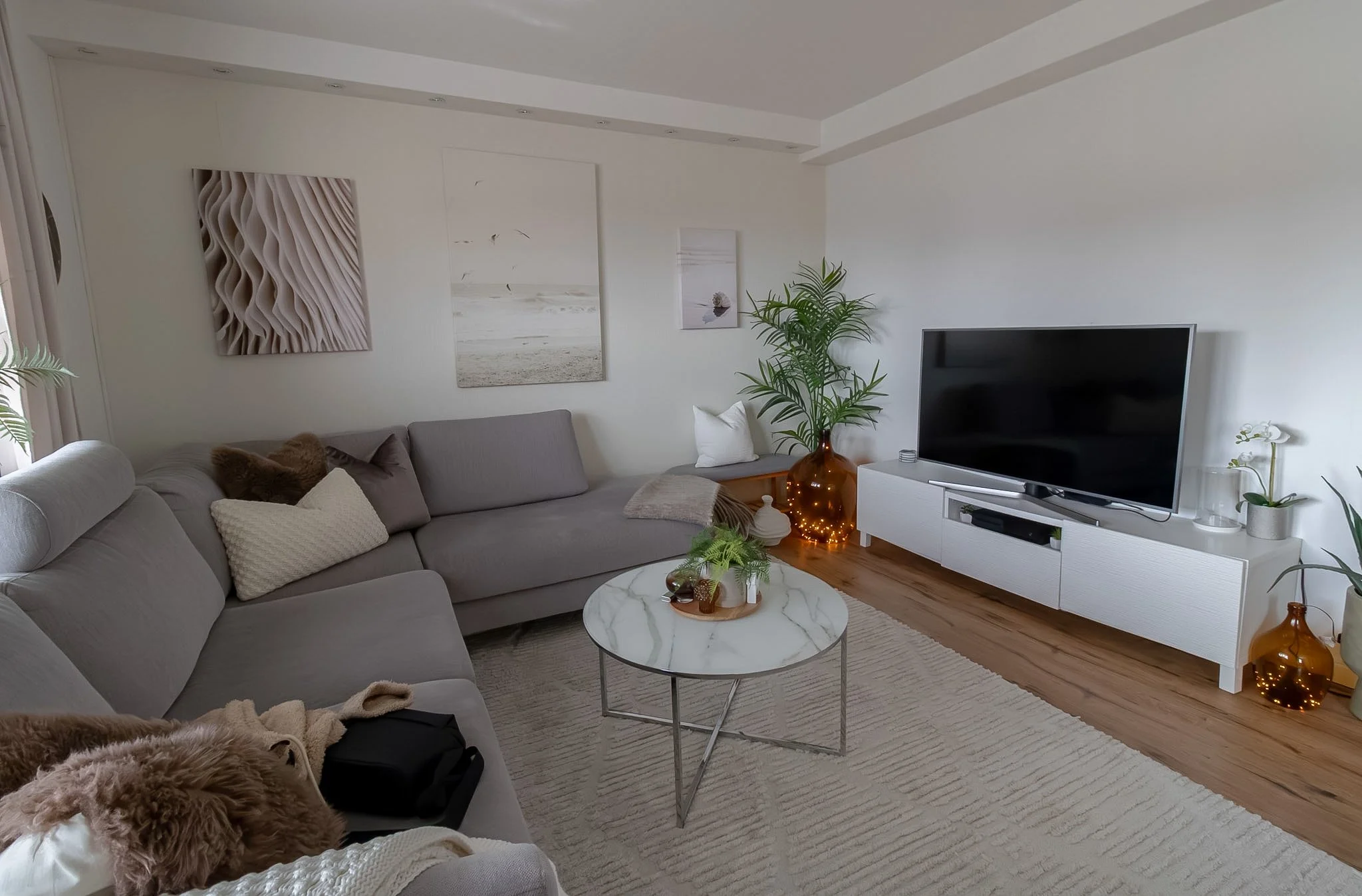 Living room with gray sectional sofa, white coffee table with marble top, wall art, potted plant, TV on white stand, and decorative vases, in a modern, minimalist interior.