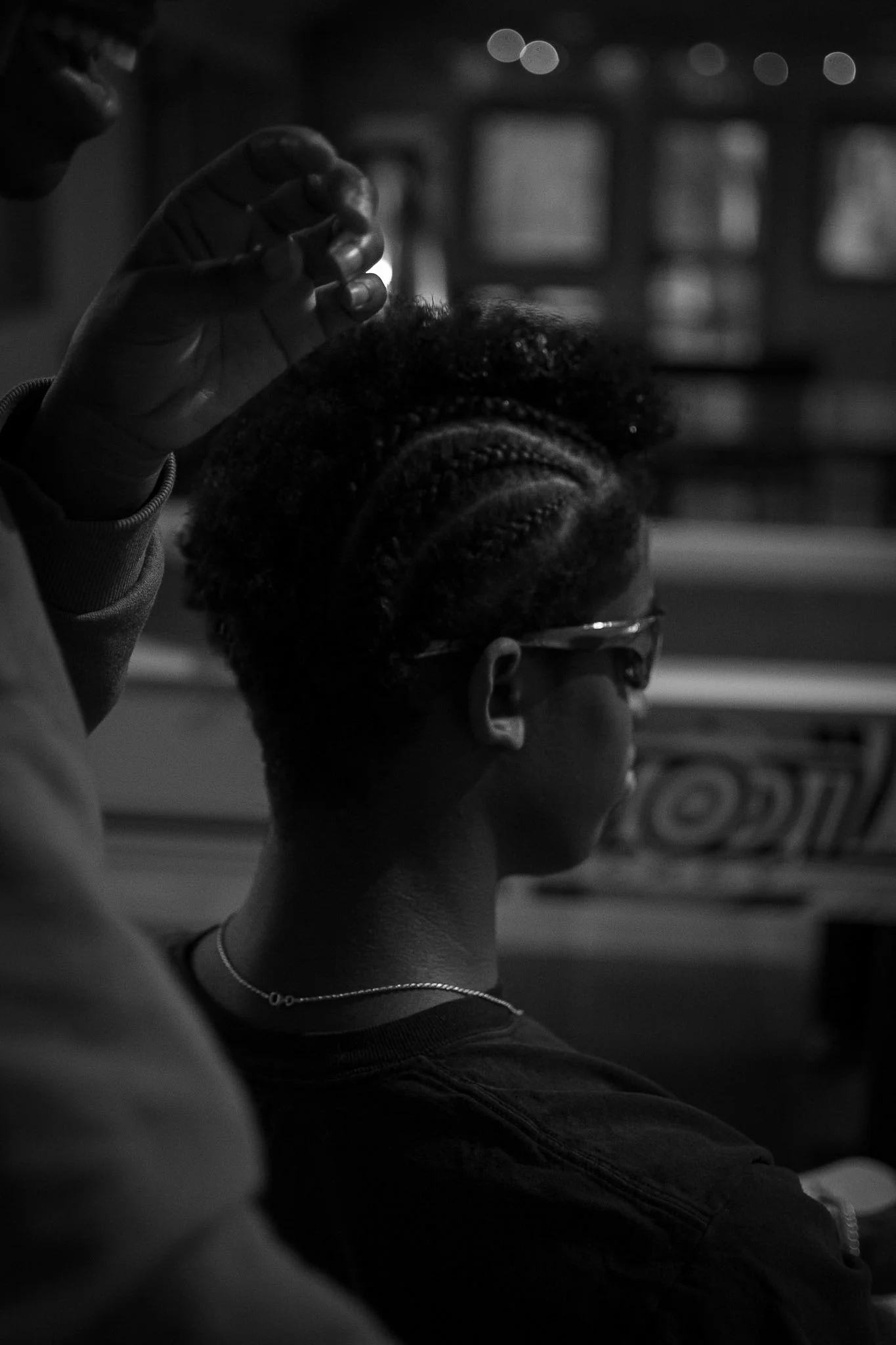 A person with curly hair wearing glasses and a necklace, sitting in a vehicle at night, with a hand of another person reaching towards their head. The background is blurry, with lights and structures visible.