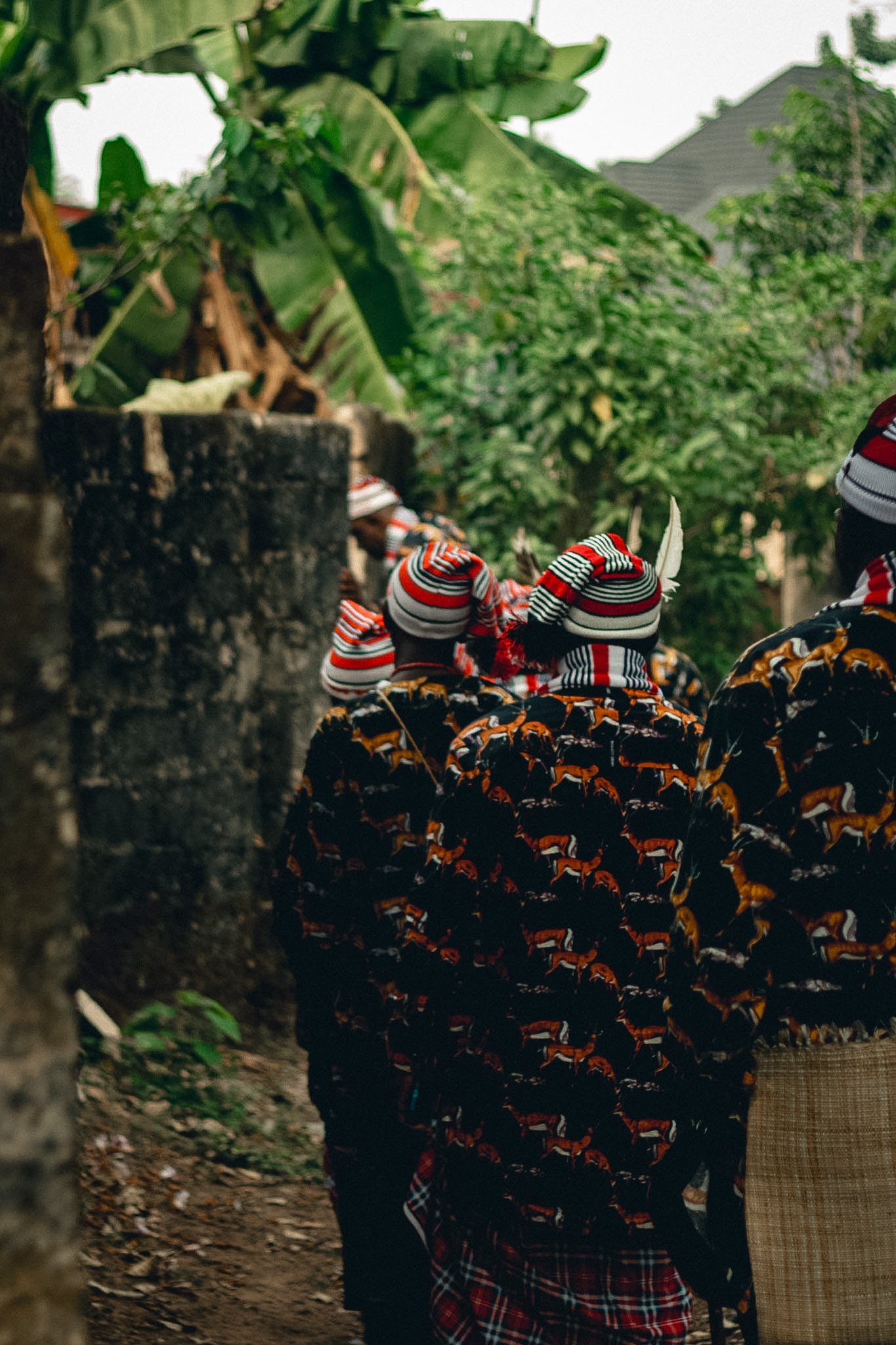 Group of people wearing traditional African clothing with colorful patterns and beanies, standing outdoors among green foliage, facing away from the camera.