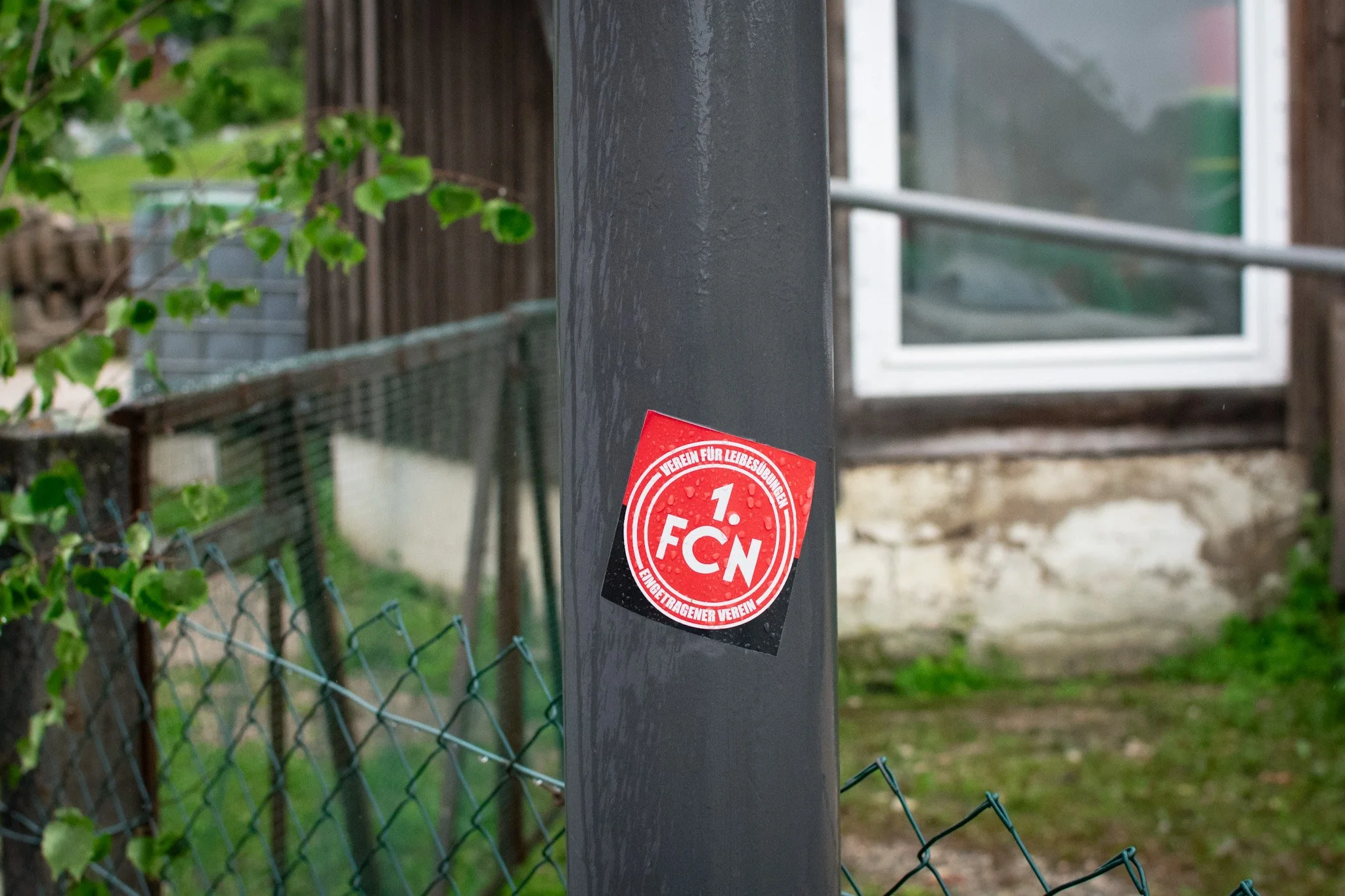 Close-up of a black metal fence post with a red circular sticker that reads '1. FCN'. Behind the post, a chain-link fence and a garden with green plants and a wooden fence are visible.
