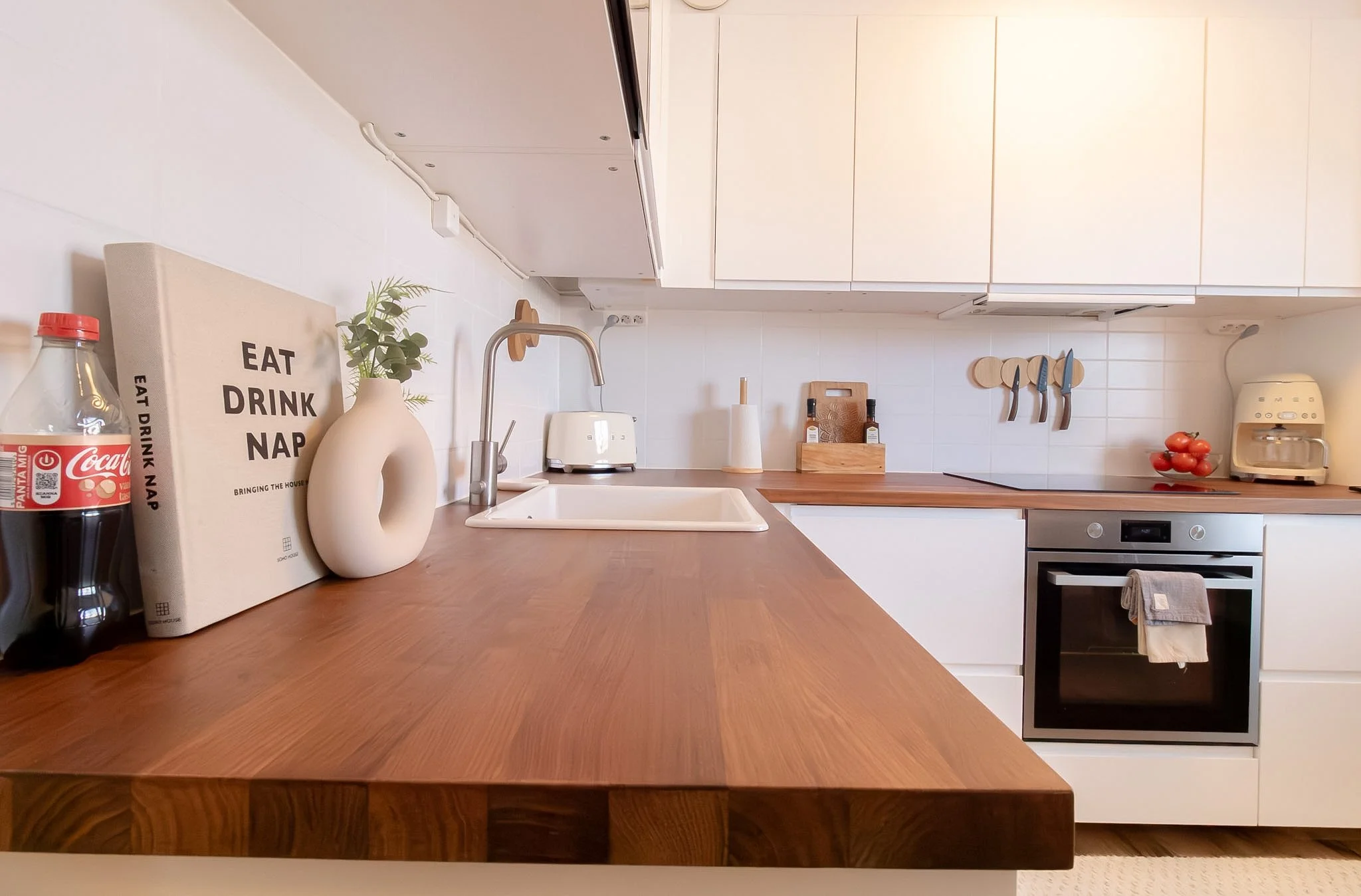Modern kitchen with white cabinets, wooden countertop, stainless steel oven, and various kitchen items including a Coca-Cola bottle, a sign that reads 'Eat Drink Nap', a small plant, a beige pitcher, a toaster, a paper towel roll, a cutting board wit
