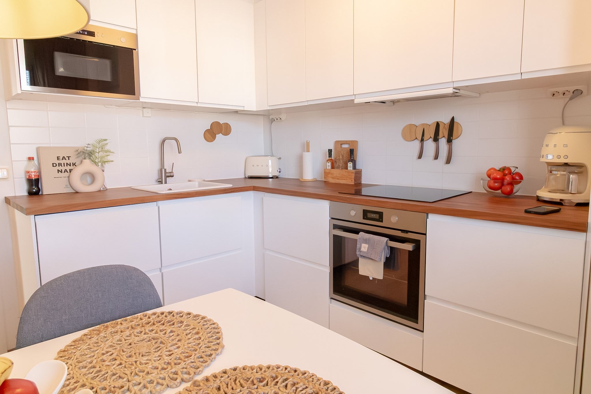 A modern kitchen with white cabinets, wooden countertops, and various appliances including a microwave, toaster, stove, and coffee maker. Decorative items like a bowl of tomatoes, cutting boards, knives, and a framed print are also visible.