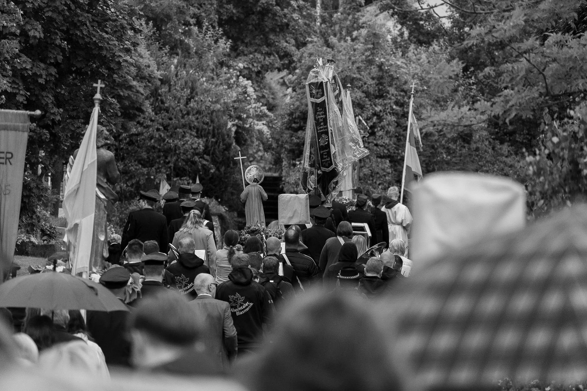 A large crowd of people gathered outdoors, facing a religious procession with crosses and flags, surrounded by trees.