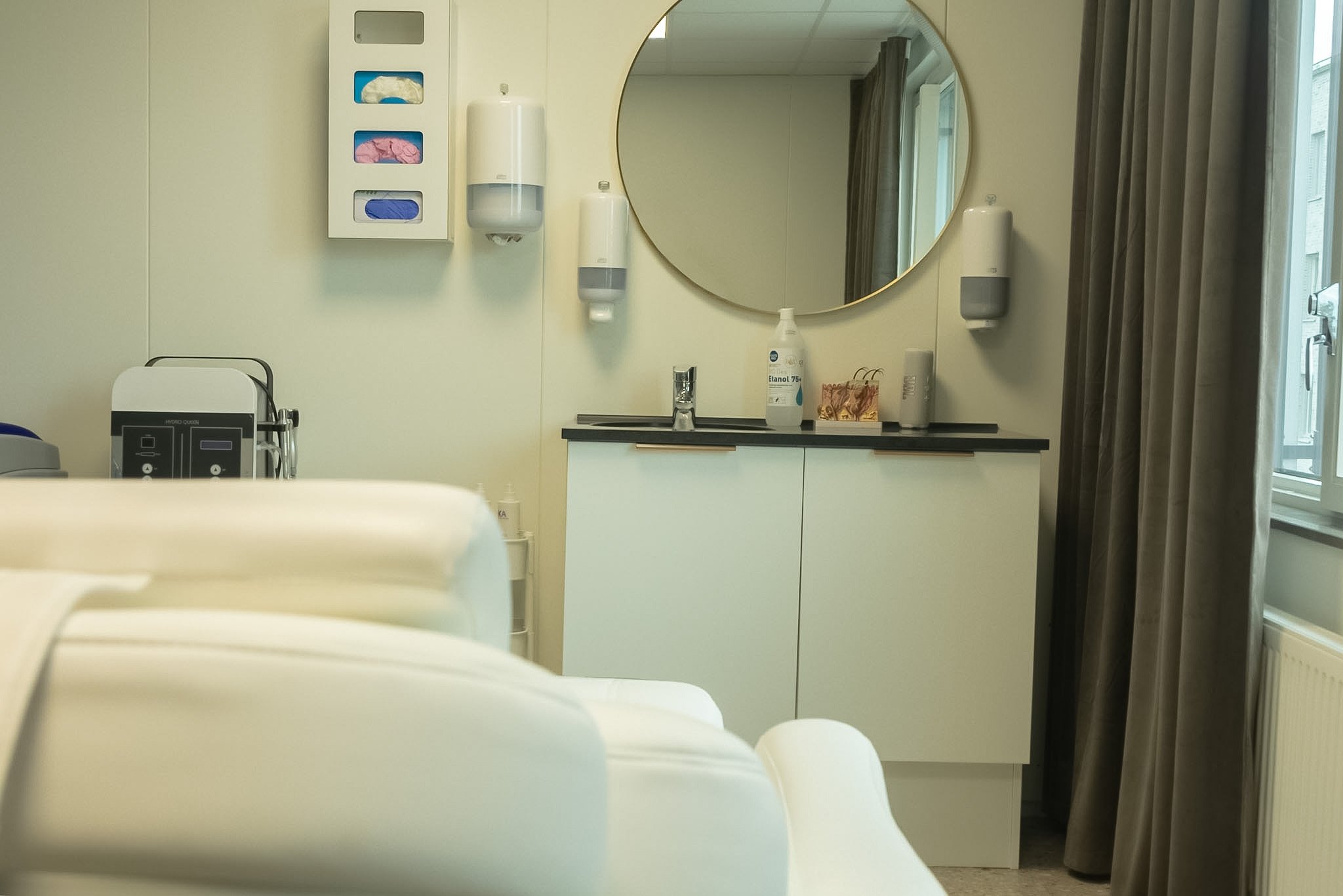 A hospital room or medical examination room with a sink, soap dispenser, sanitizer, and a large mirror on the wall. Curtains cover windows, and medical equipment is visible in the foreground.