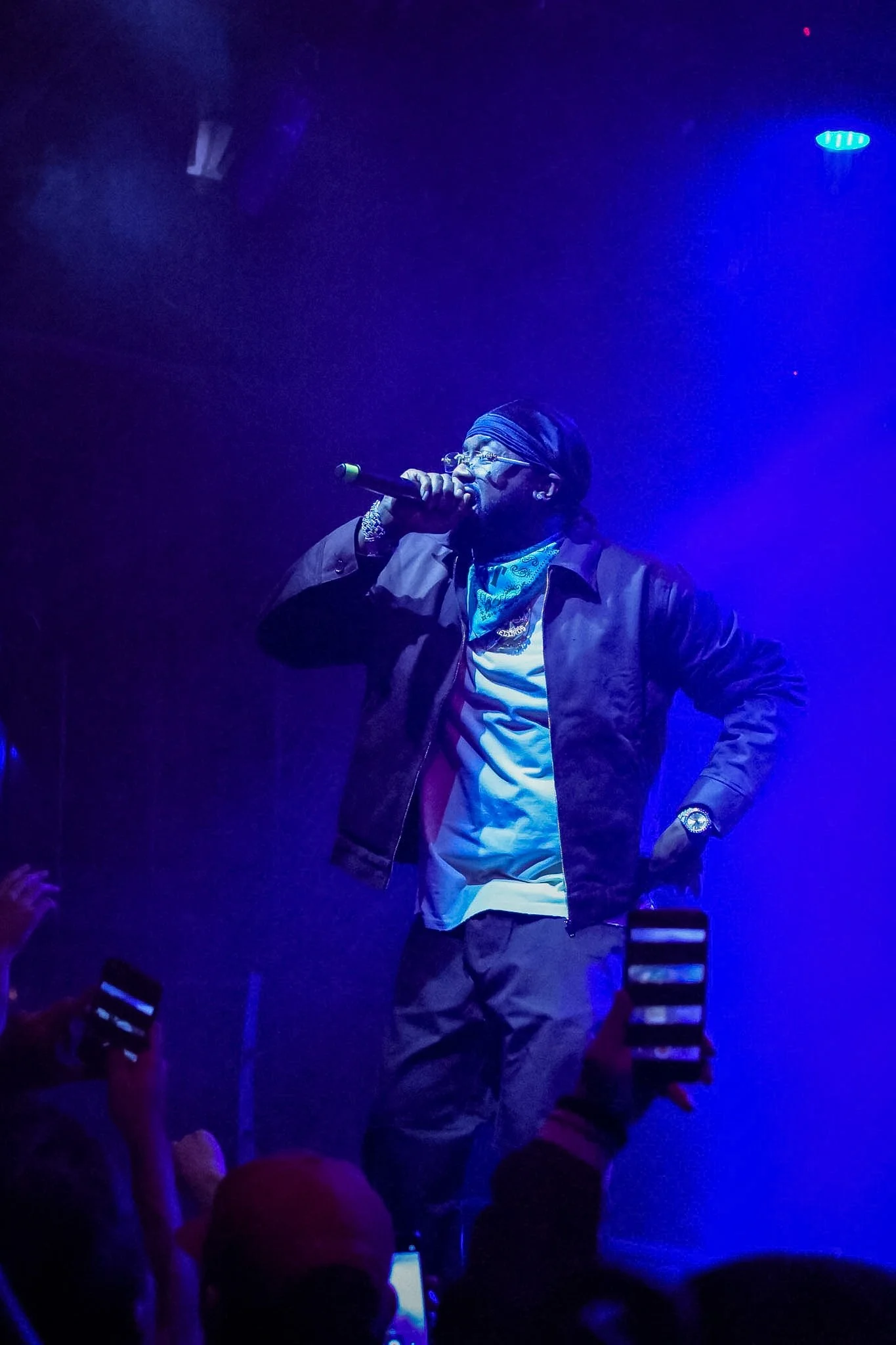 Male performer with glasses and a headscarf singing into a microphone on stage, lit by blue lighting, with audience members capturing the moment on their phones.