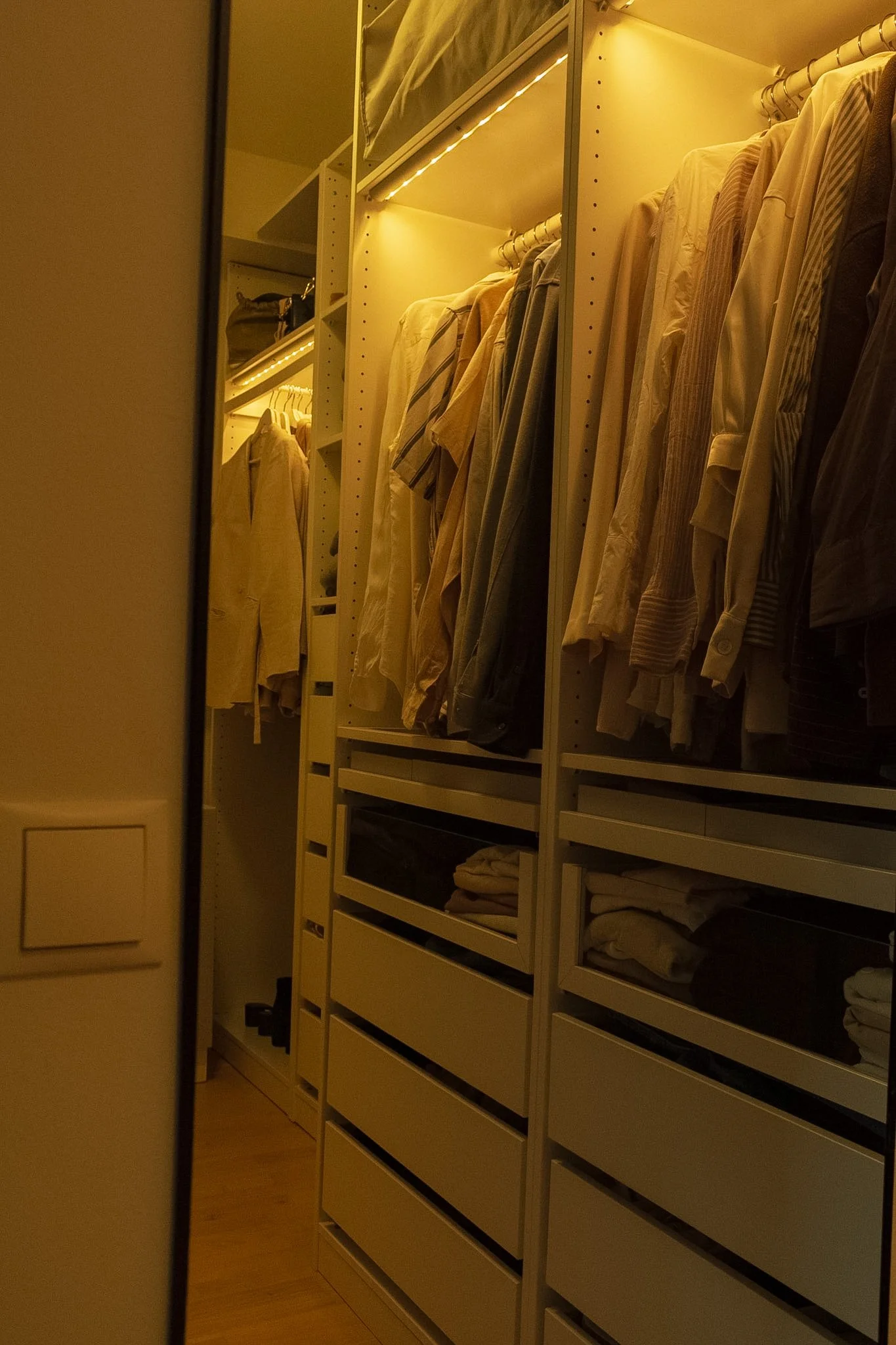 A walk-in closet with white shelves and hanging clothes illuminated by warm lighting.