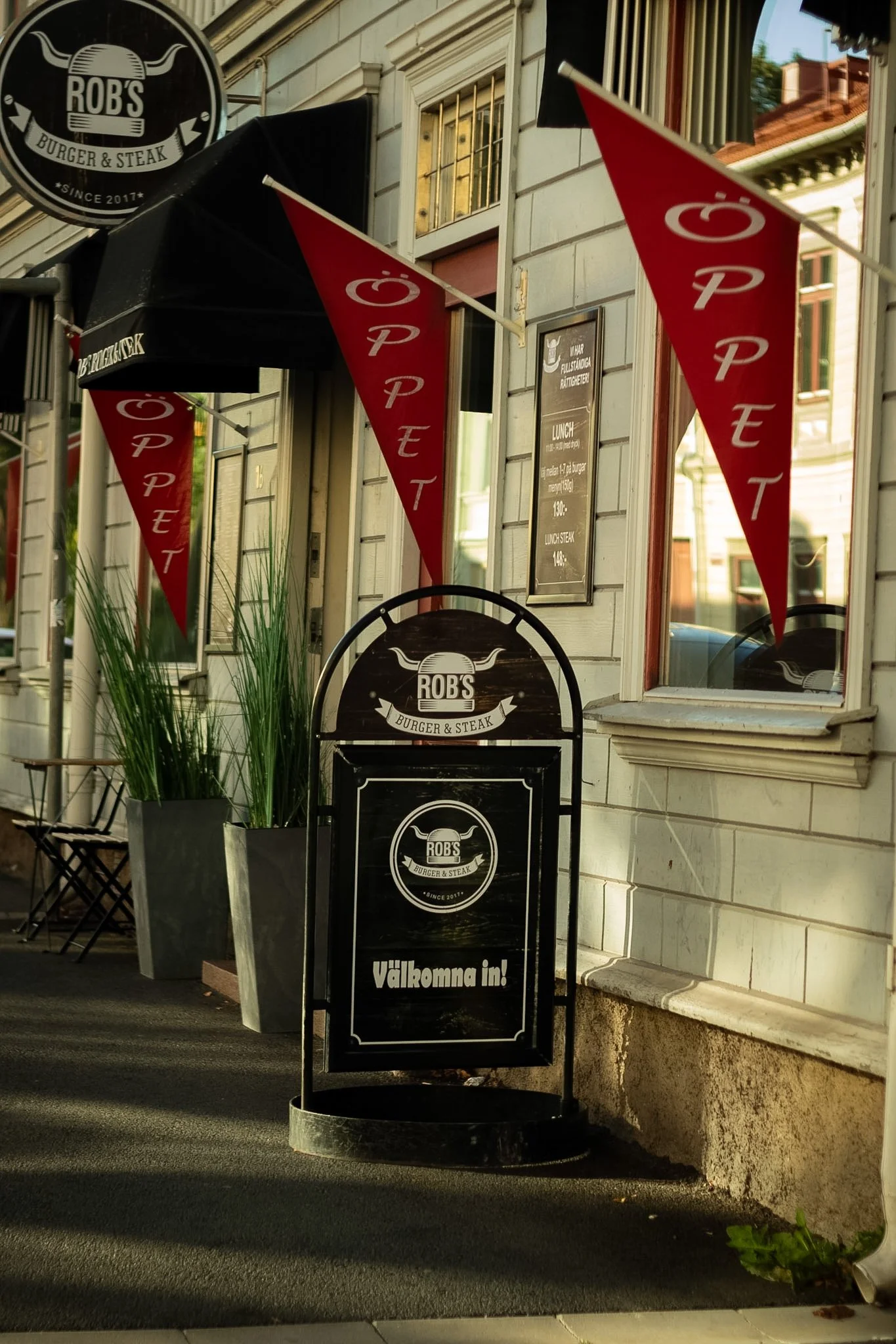 Exterior of a restaurant called Rob's Burger & Steak with black and red flags, and a black sandwich board sign that says "Välkomna in!" (Welcome in!) in Swedish. The building has white wooden siding, a window, and a black awning with the restaurant's