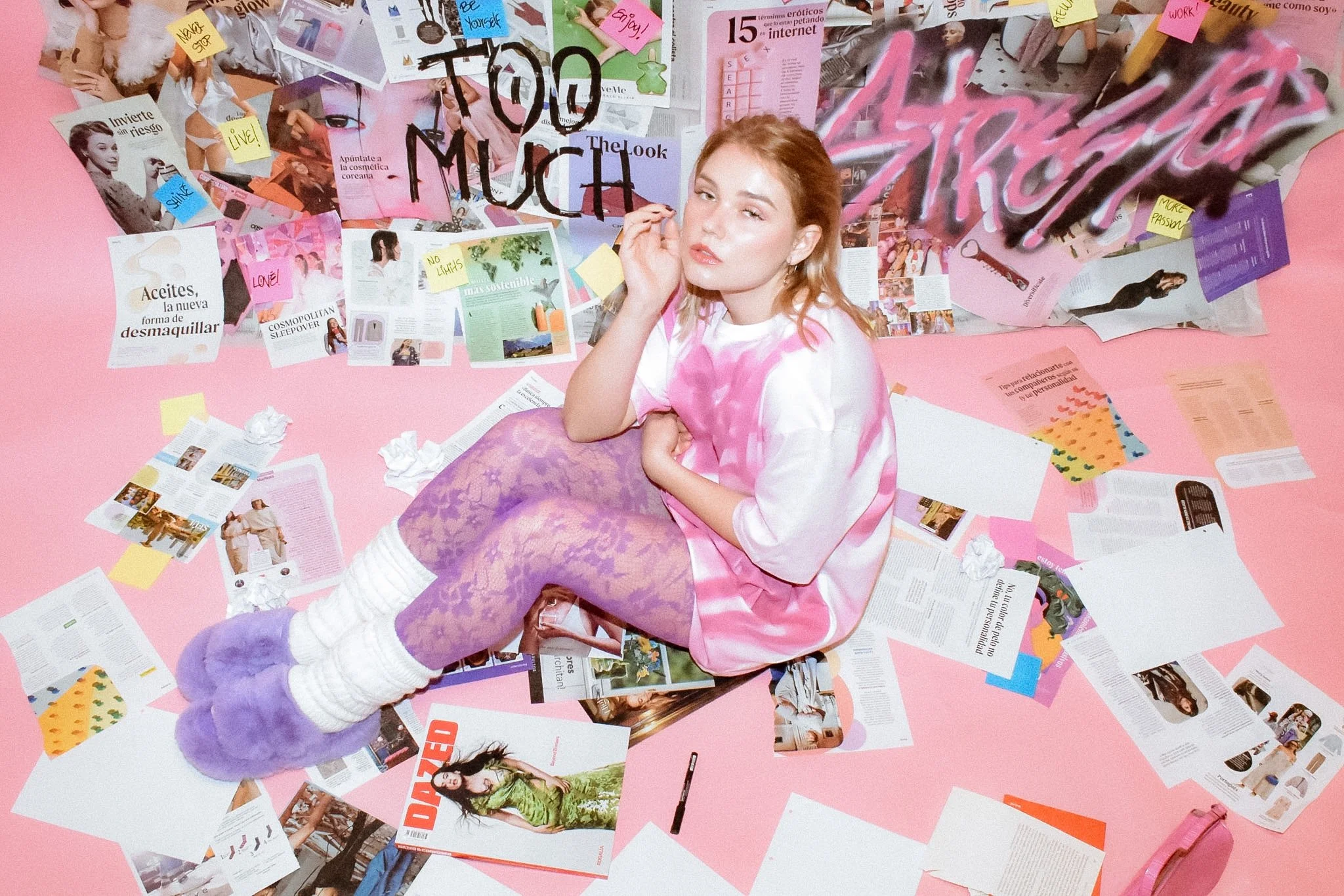 A young woman with blonde hair sitting on a pink floor, surrounded by magazines, papers, and sticky notes, with her head tilted and hand touching her face.