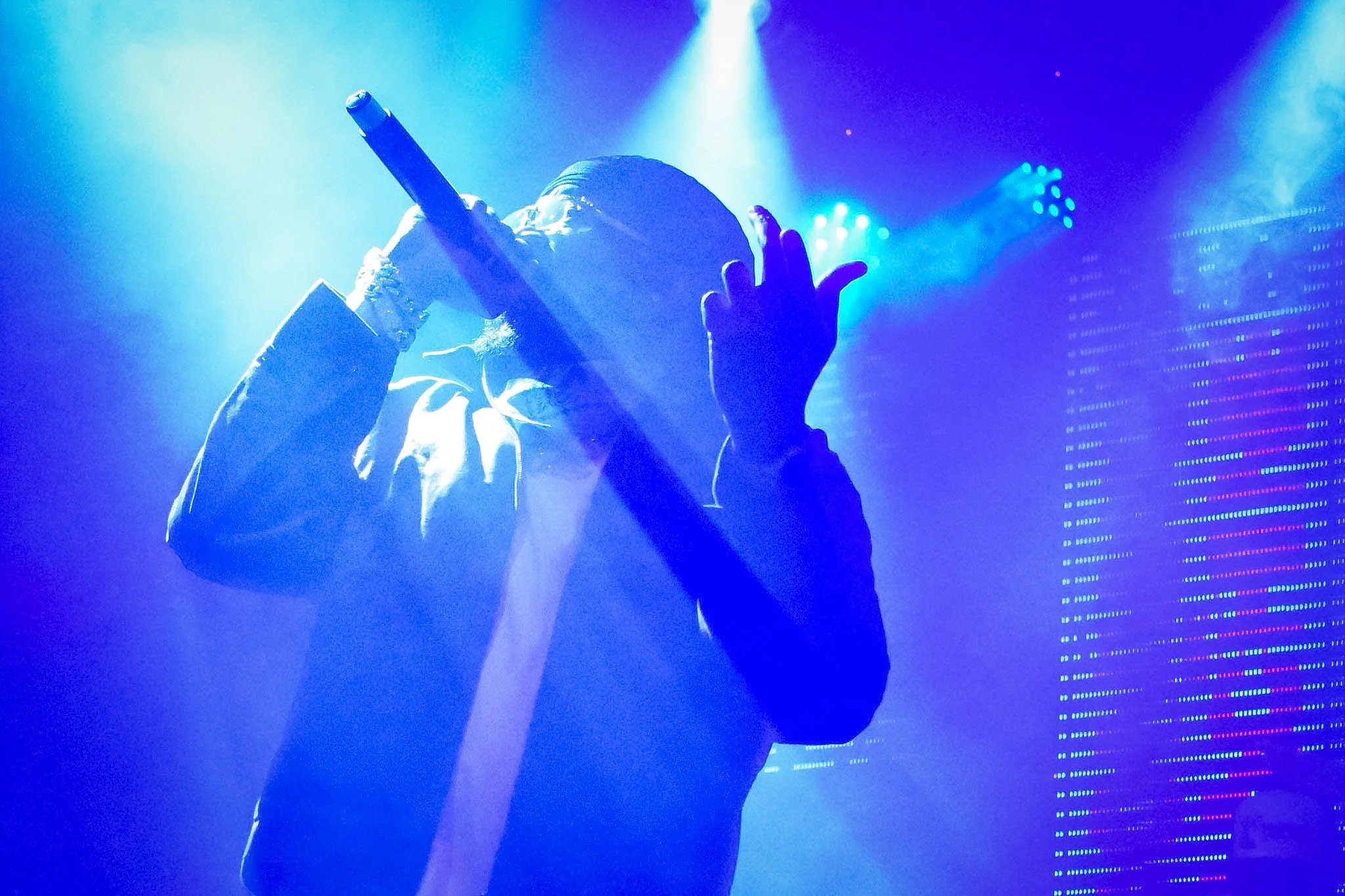 Performer singing into a microphone on stage illuminated by blue and purple lights with an LED display in the background.