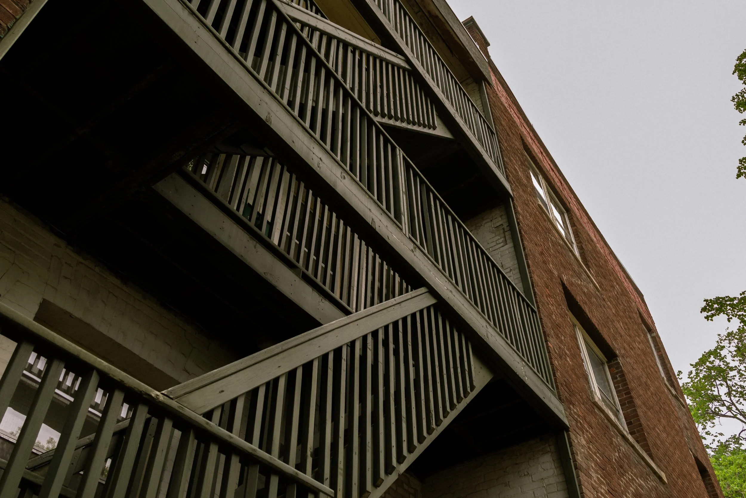 A close-up view of an apartment building with multiple balconies featuring wooden railings and brick exterior walls, with a cloudy sky in the background.