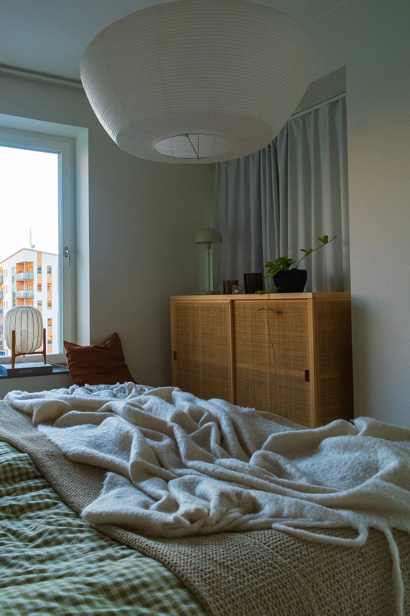 A cozy bedroom with a bed covered in a white, fuzzy blanket, a brown pillow, and a woven textile. Near a window, there is a wooden side table with a lamp and a decorative paper lantern. A wooden cabinet with a potted plant on top is against the wall.