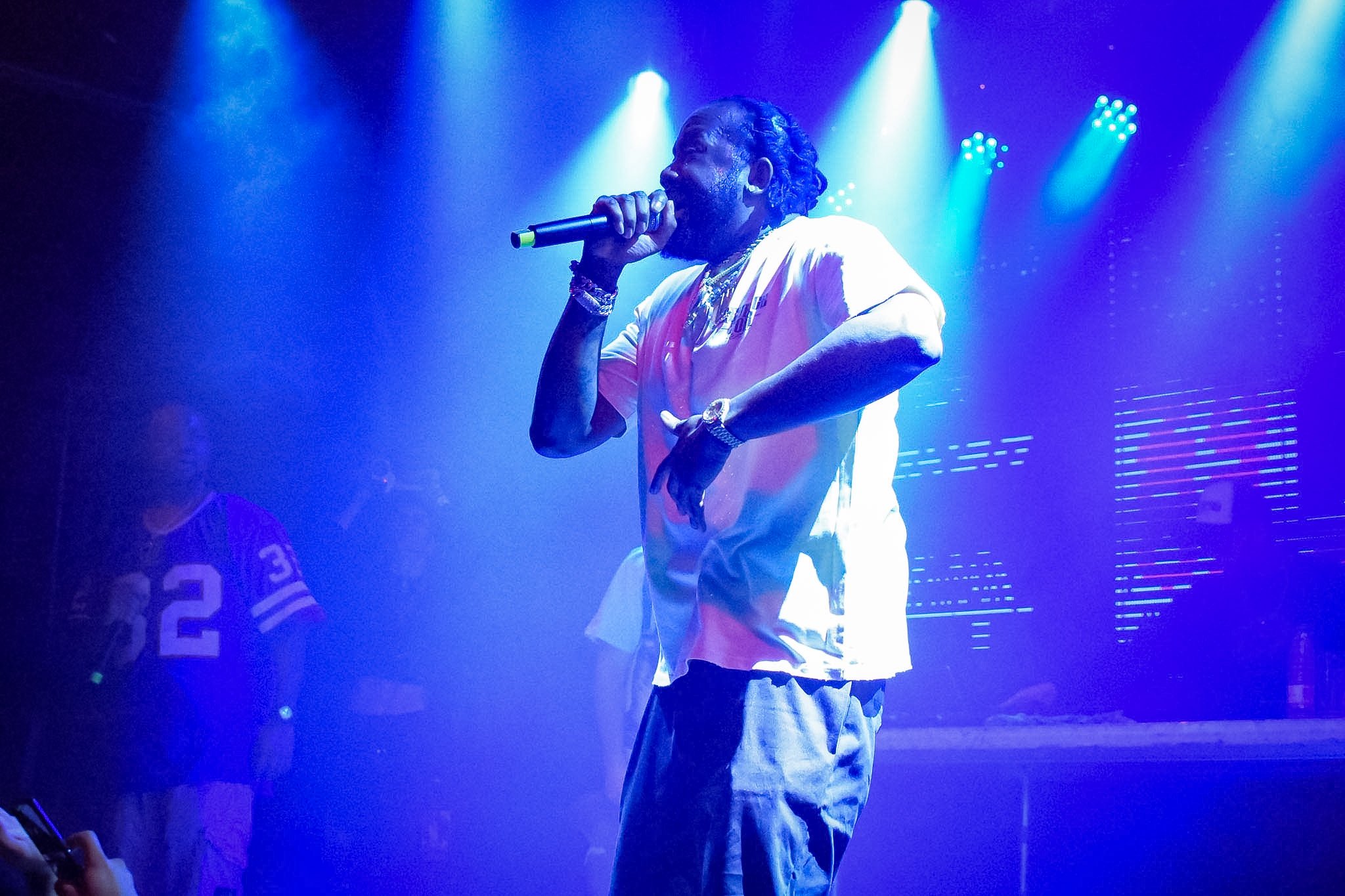 A male performer with long, braided hair singing into a microphone on stage, with blue stage lighting and a person wearing a sports jersey visible in the background.