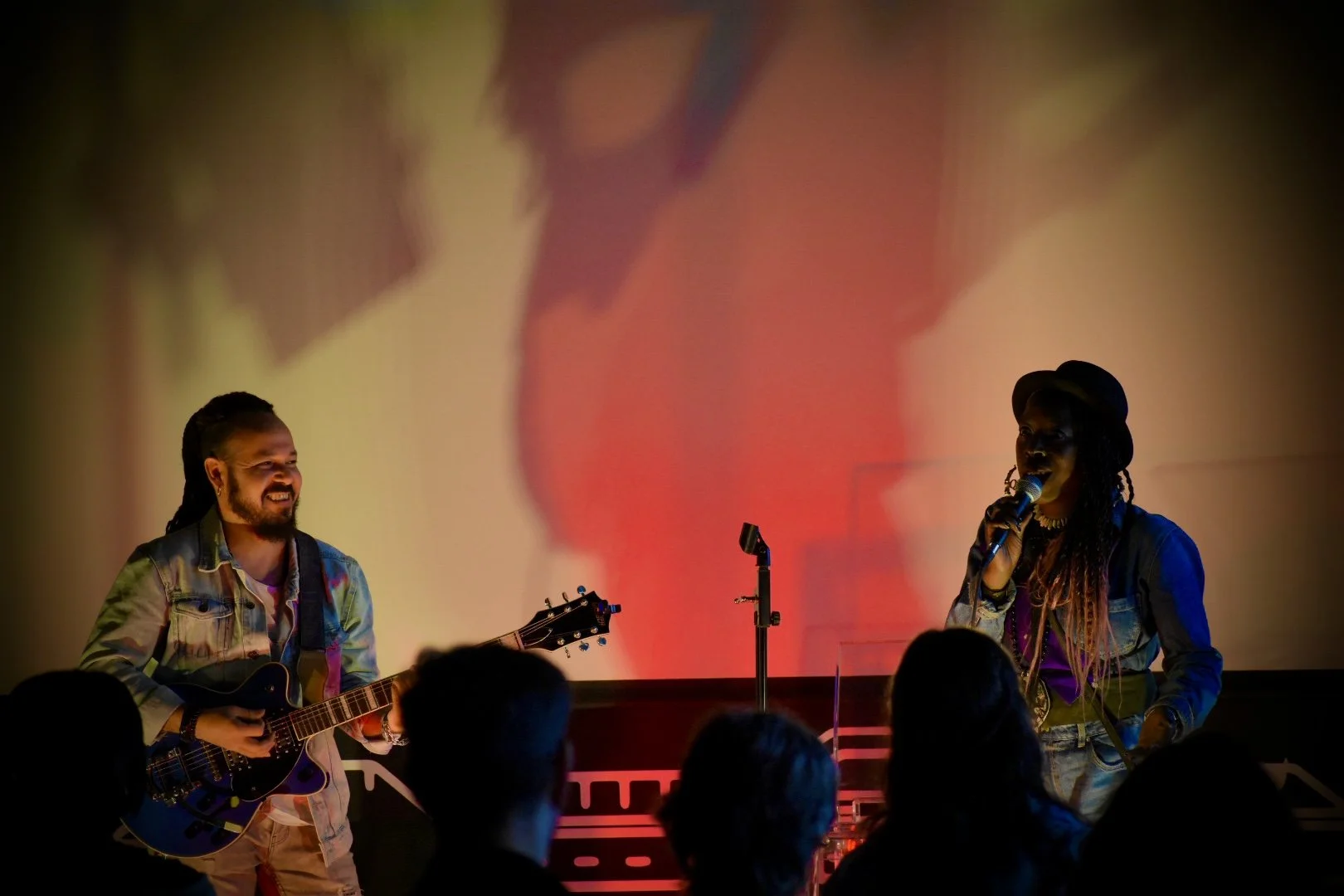 Musicians performing on stage with a guitar and microphone, with an audience watching, illuminated by warm lighting and a large screen projecting abstract visuals in the background.