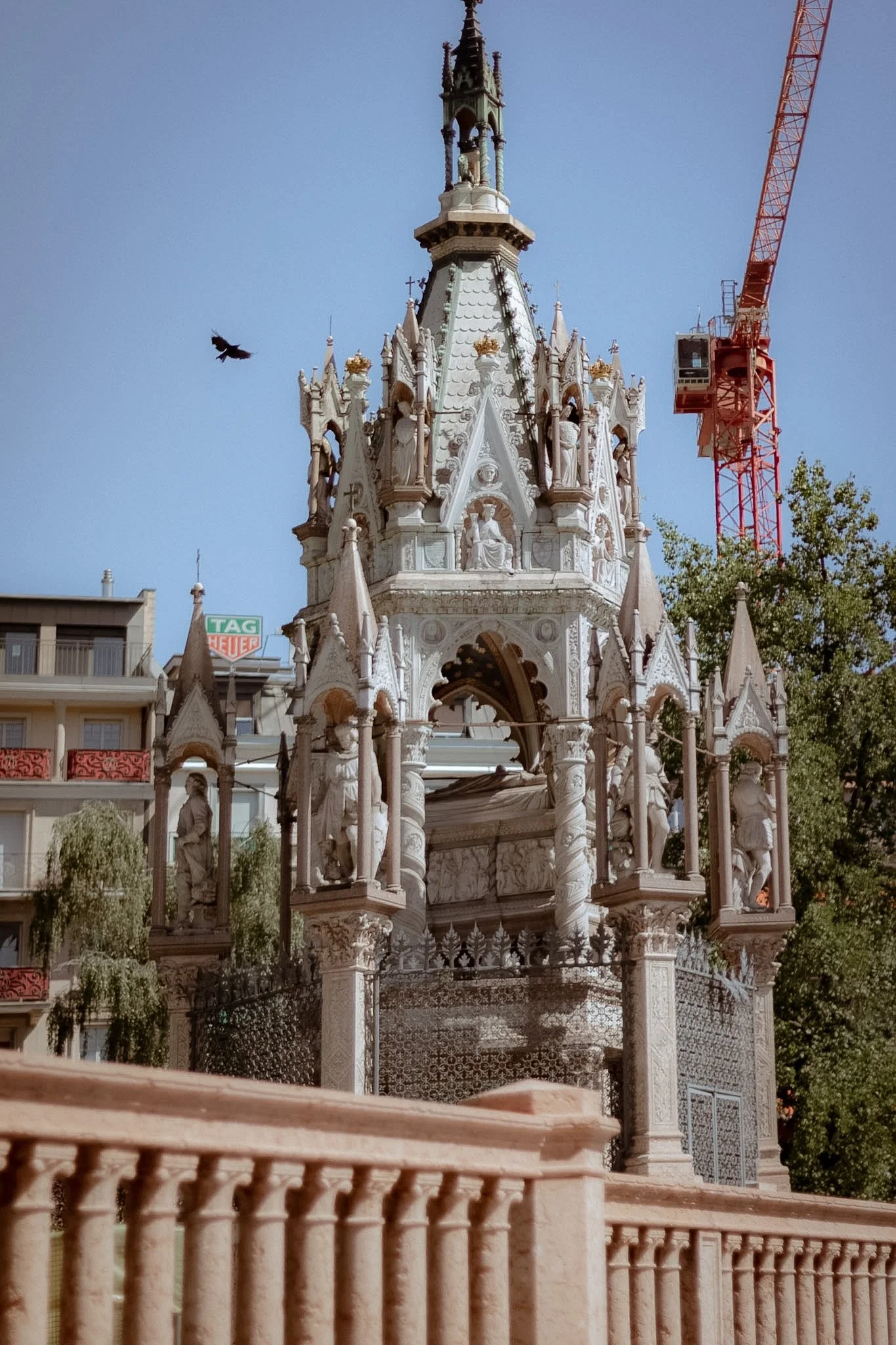 A detailed white ornate structure with statues and Gothic architectural elements, topped with a spire, in an urban setting with buildings and trees nearby, and a red crane visible in the background.