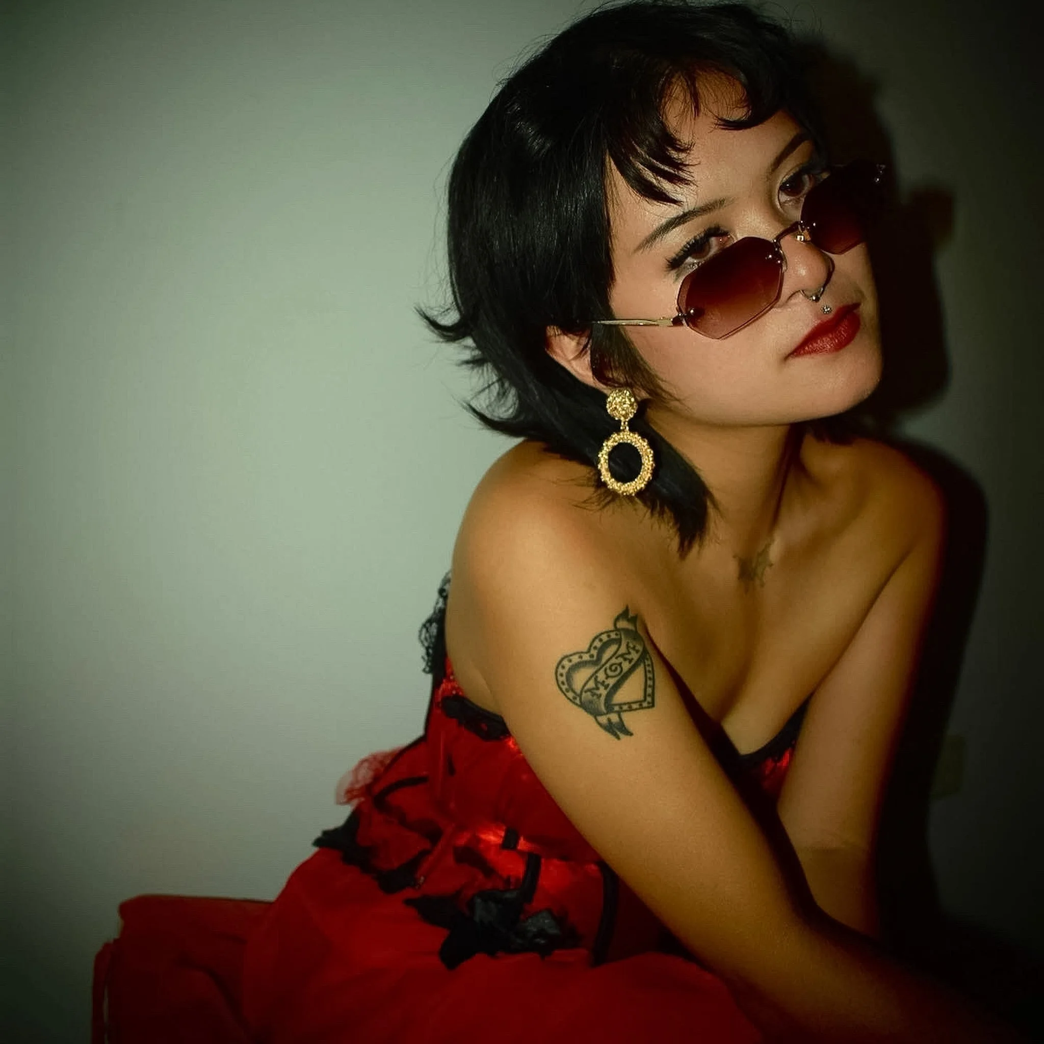 A woman with short black hair wearing red glasses, red lipstick, gold earrings, and a black tattoo on her upper arm, dressed in red and black lace lingerie, looking over her shoulder against a plain background.