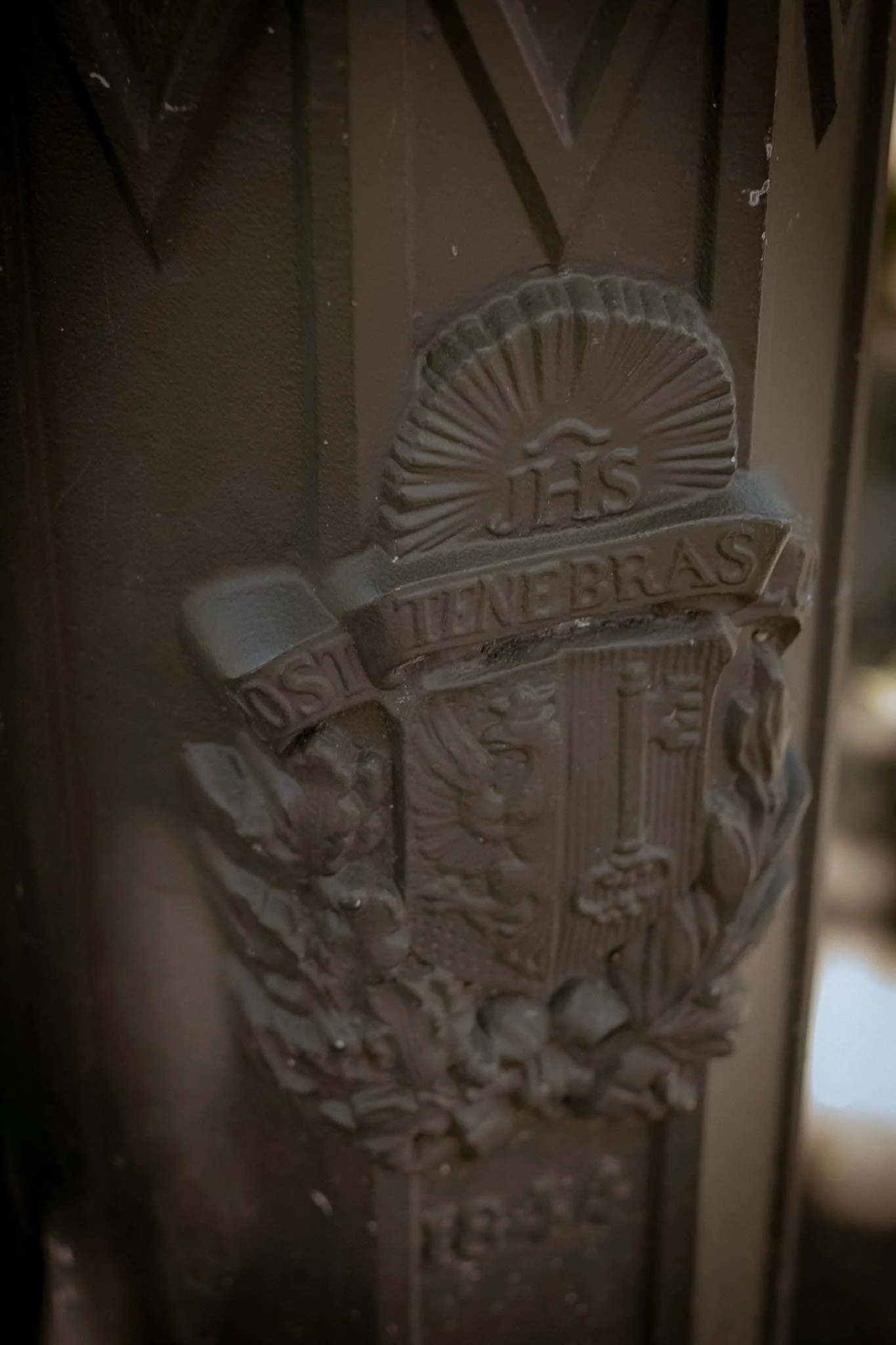 Close-up of a shield-shaped emblem with the inscription 'JHS' and the word 'TANNER' on it, mounted on a textured surface.