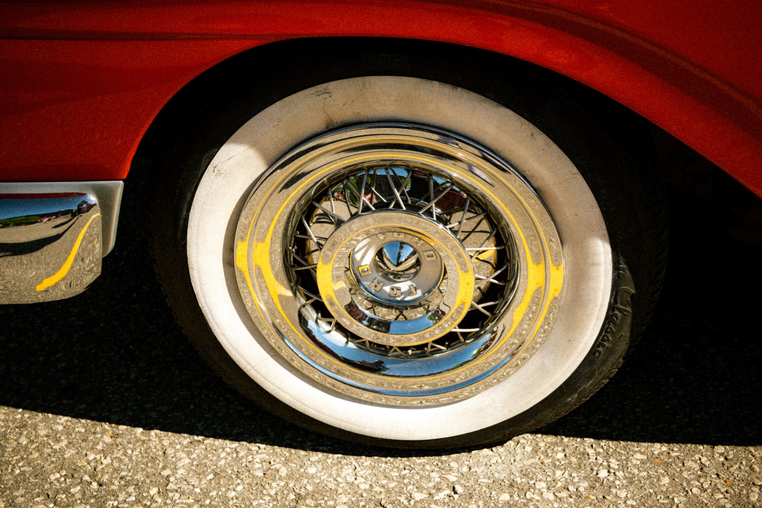 Close-up of a vintage car wheel with a wire-spoke rim and white-wall tire, parked on an asphalt surface.