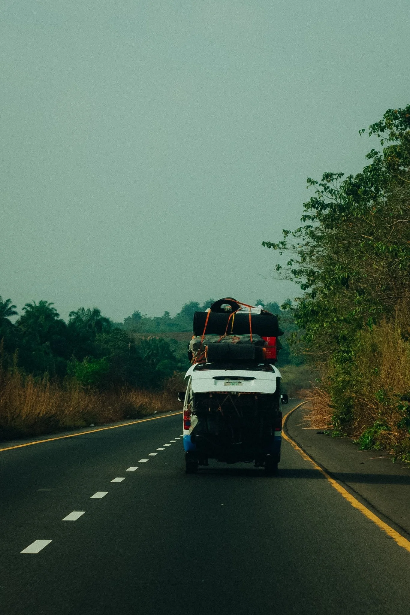 Two vehicles driving on a highway with trees on either side. The vehicles are carrying large loads with luggage and gear tied on top.