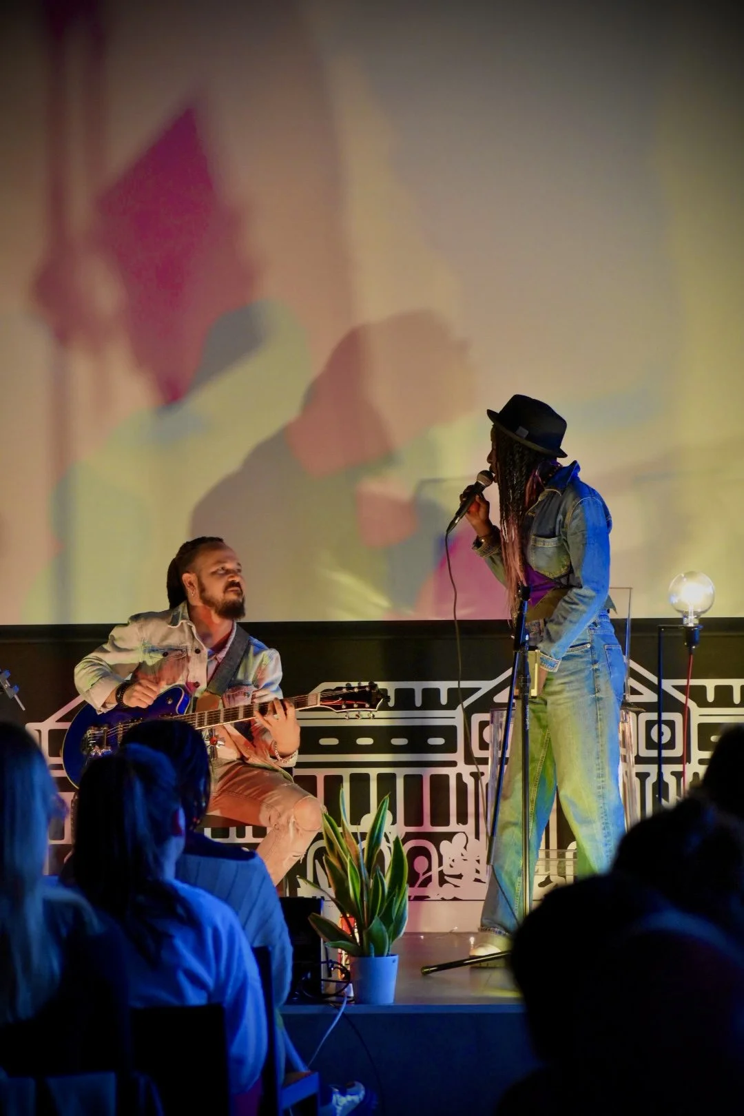 A musical performance with a male guitarist sitting and playing, and a female singer standing with a microphone on stage. The stage has a large projected colorful background, and audience members are visible in the foreground.