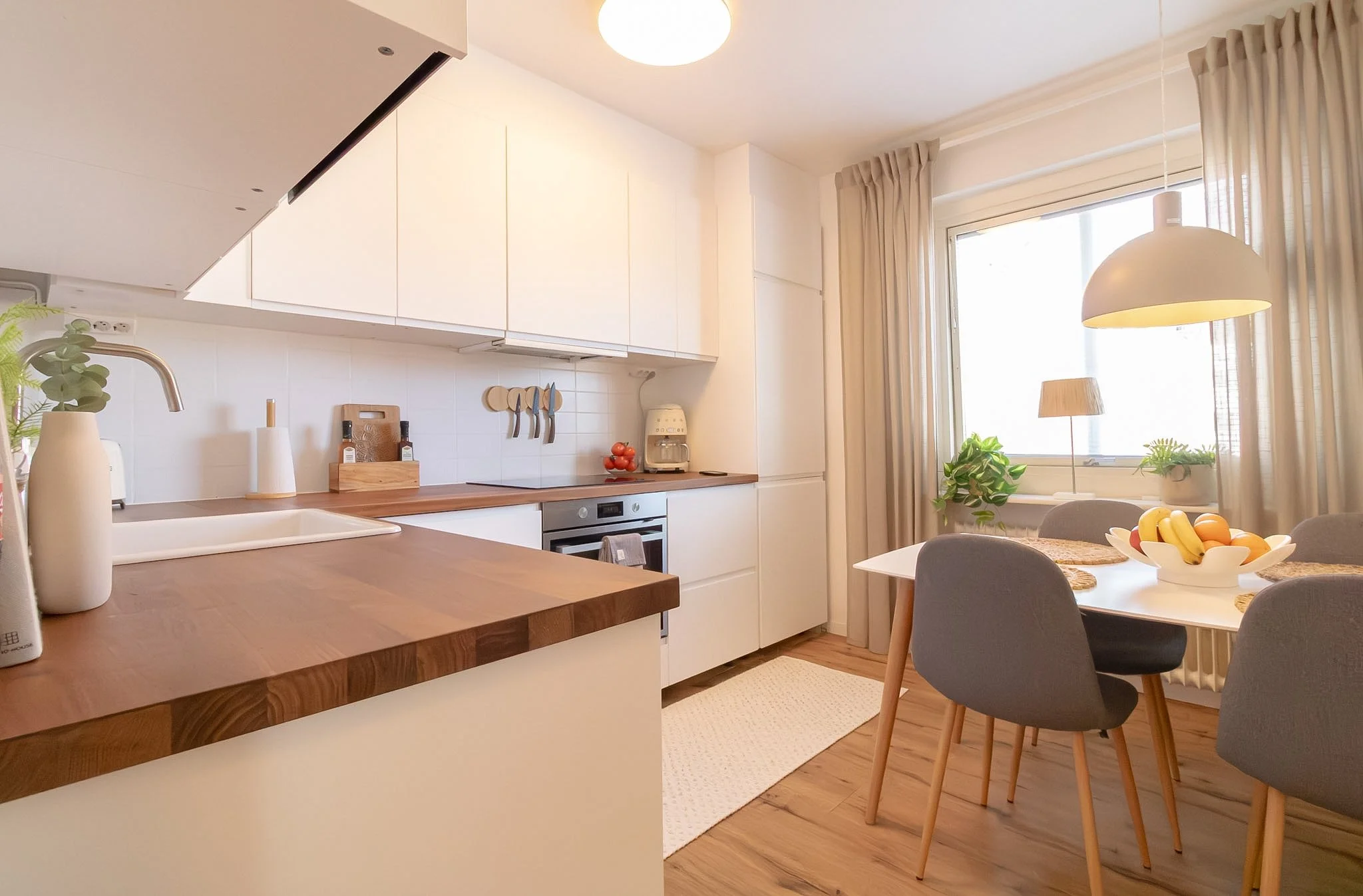 Bright modern kitchen and dining area with white cabinets, wooden countertops, and a window with beige curtains. The dining table has a fruit bowl with bananas, oranges, and apples, and gray chairs around it.