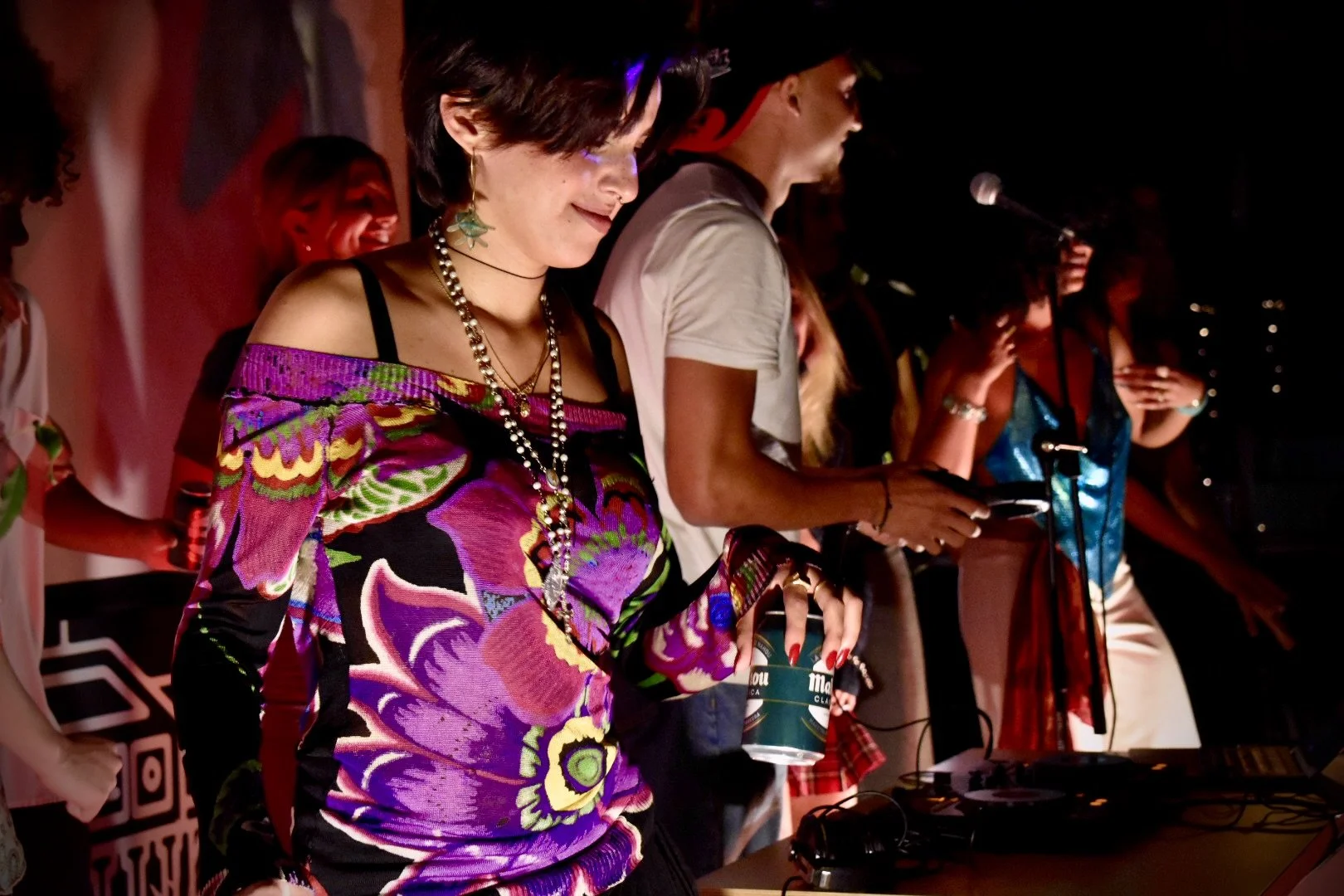 Group of people enjoying a nighttime party or event, some using DJ equipment and mobile phones, with one person in colorful floral attire and jewelry in the foreground.