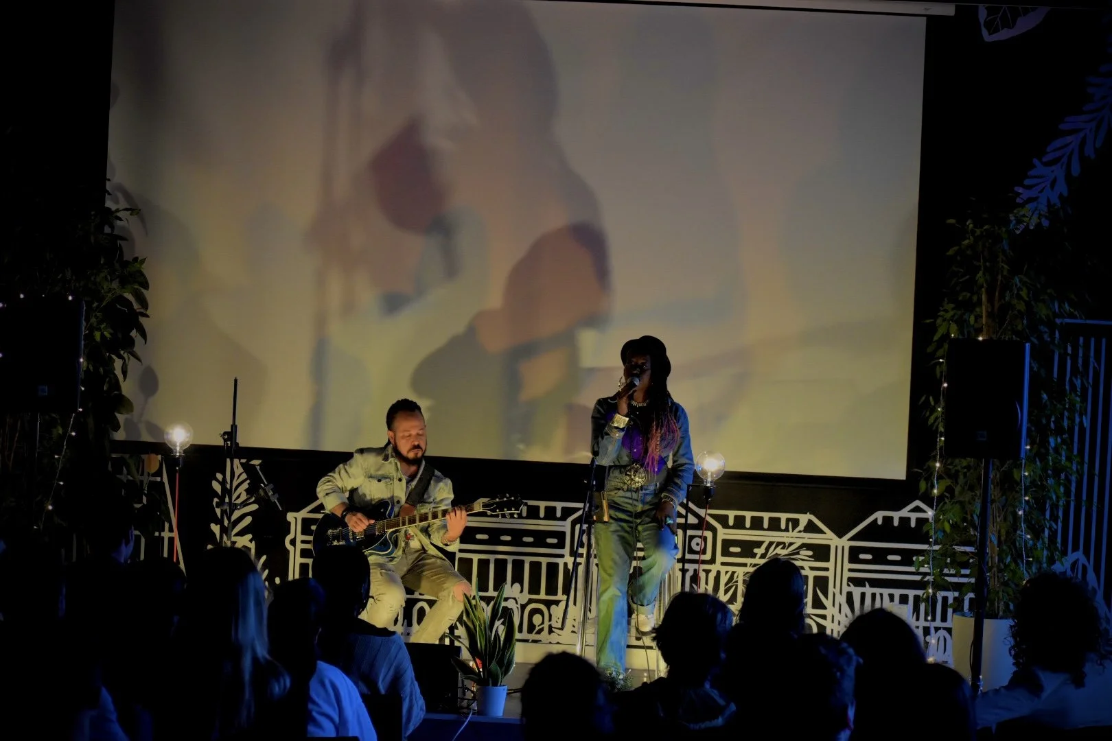 A live musical performance on a stage with a woman singing into a microphone and a man playing an electric guitar. The stage features decorative elements, plants, and a large screen projecting a blurred image of the performers.