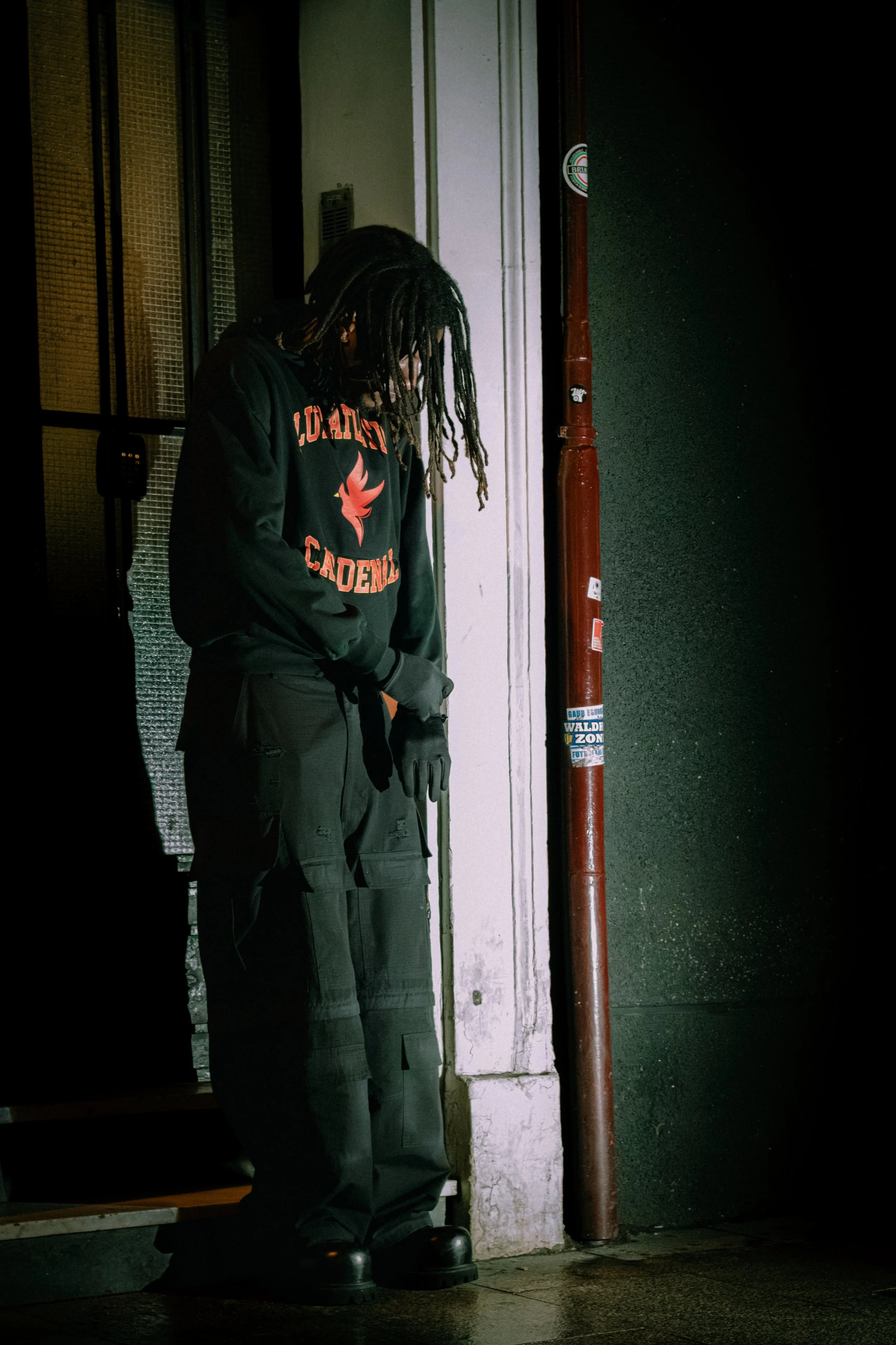A person with dreadlocks and glasses wearing a hoodie with the words 'Lionsgate Academy' and a lion mascot, standing against a wall near a red pipe and door, in a dimly lit area.