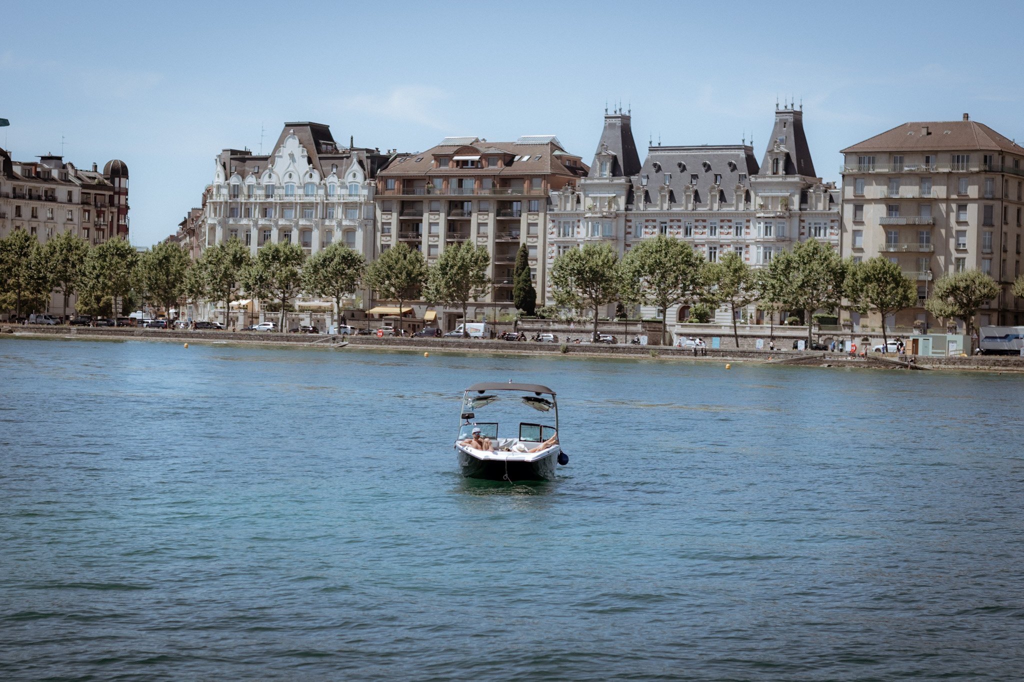 A boat on the water with a cityscape of European-style buildings along the riverbank, trees lining the street, and a clear sky.