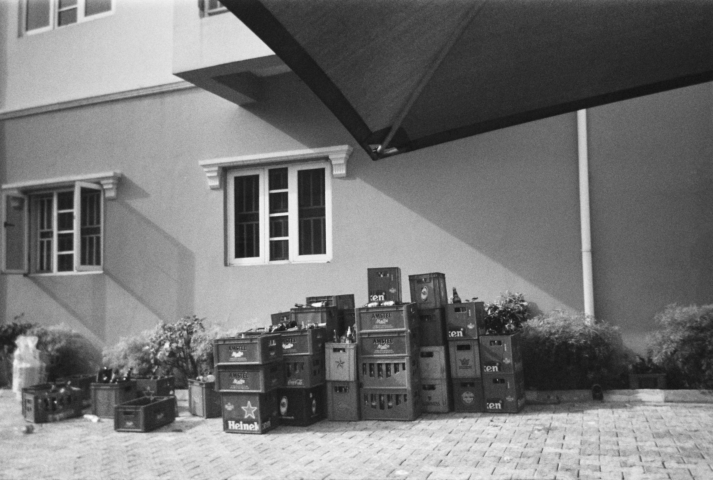 Black and white photo of a residential building with two visible windows, boxes with drinks stacked outside, and a canopy extending from the building.