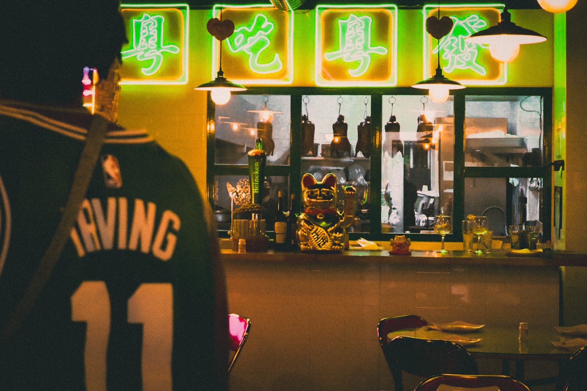 Night scene at a bar or restaurant with neon sign writing in Chinese characters, lit pendant lights, a small Golden Buddha statue on the counter, and a person wearing a dark shirt with an orange and white design on it, sitting or standing at the coun