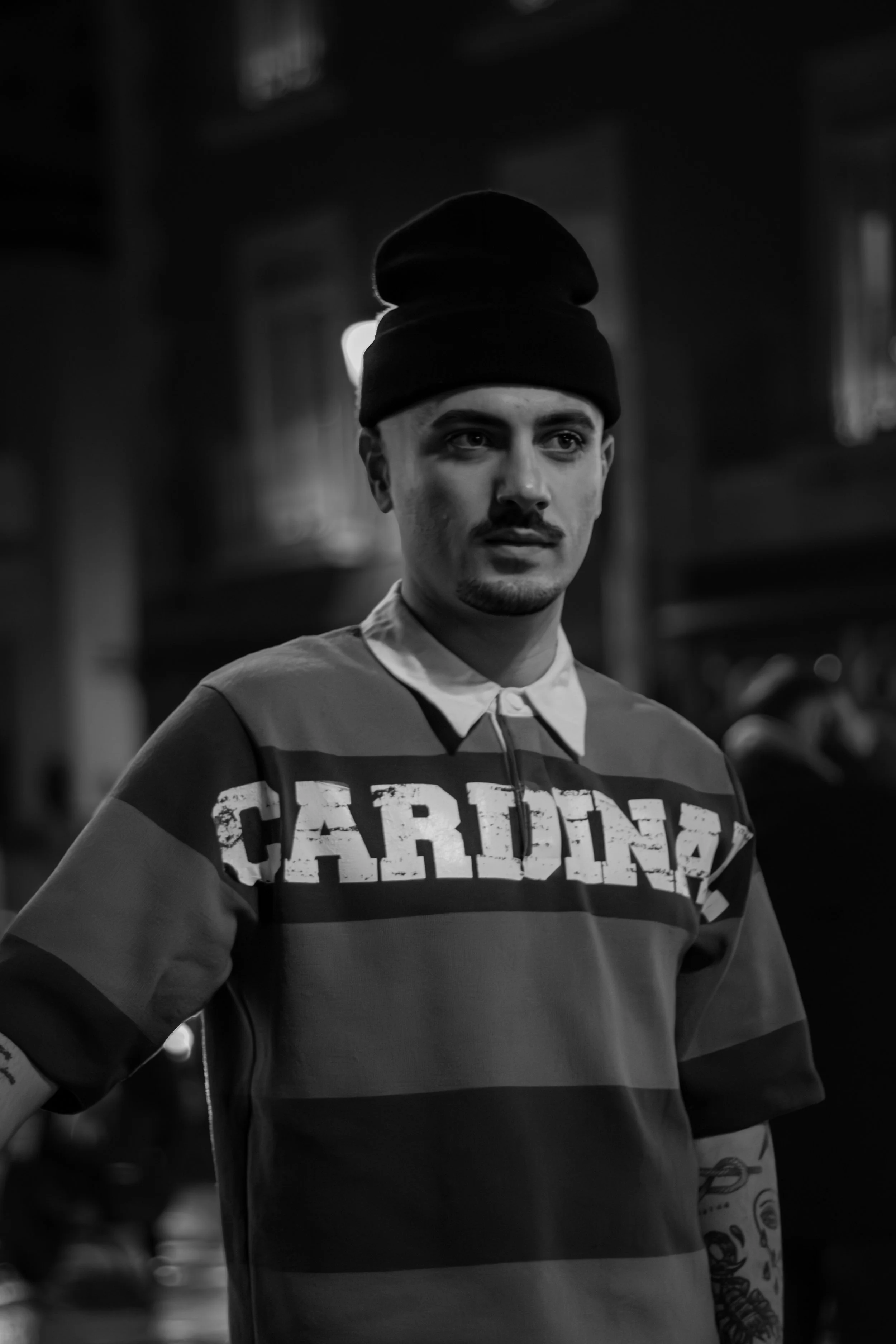 A young man wearing a striped sweatshirt with the word "CARTEL" on it and a black beanie, in a black and white photo.
