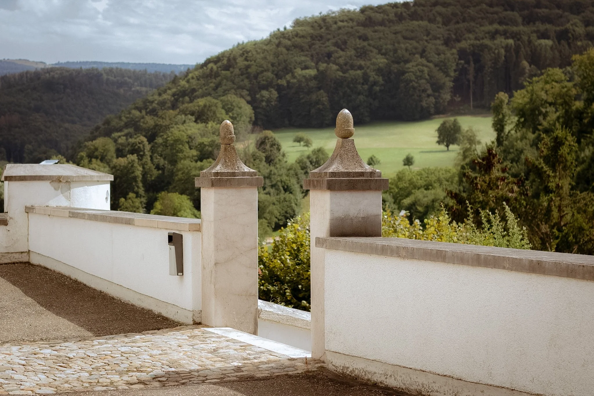 A scenic view of a countryside landscape with green rolling hills and trees, seen from a balcony with white stone posts topped with decorative stone finials, against a cloudy sky.
