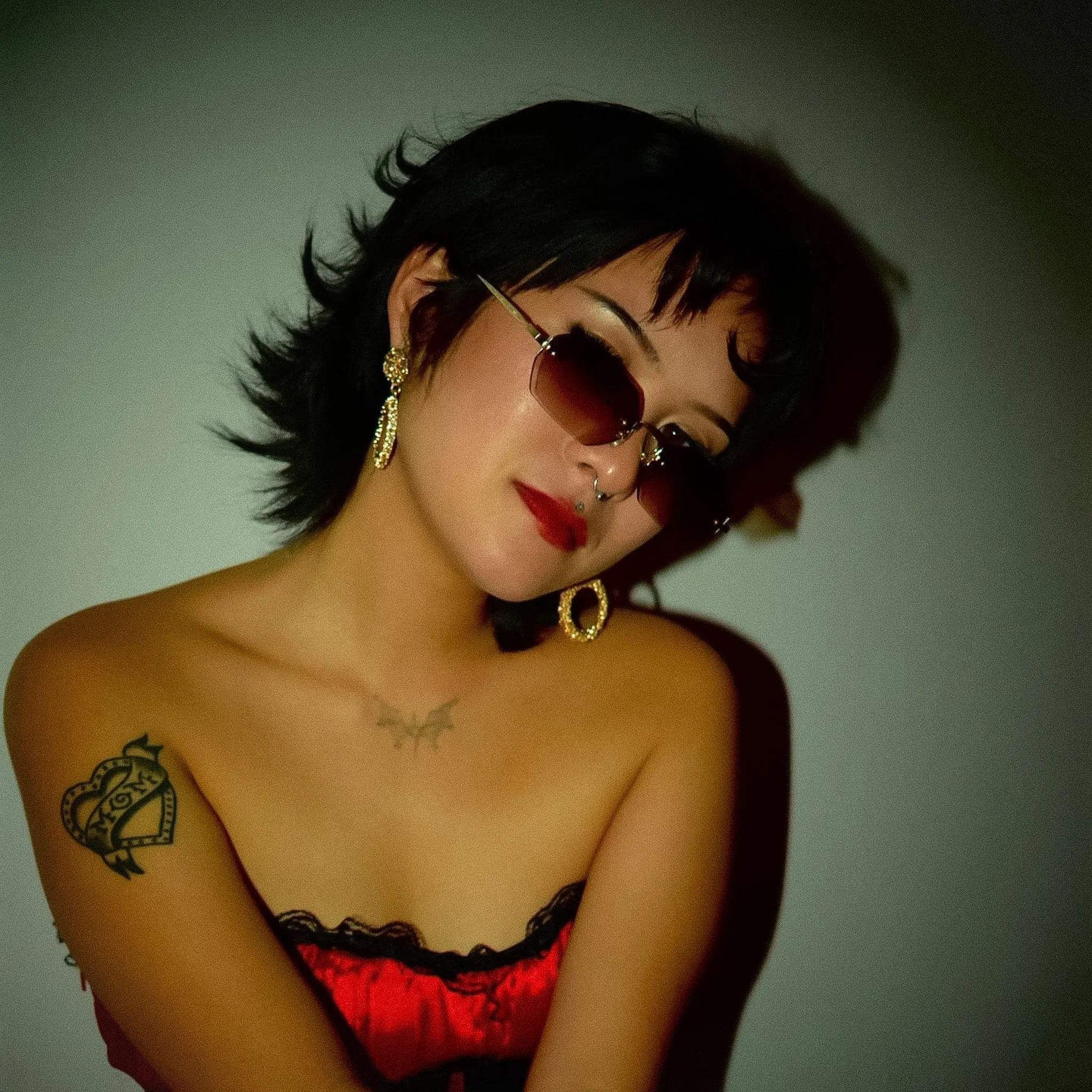 A woman wearing a red lace bra, sunglasses, and jewelry with tattoos on her arm and neck, posing against a plain background.
