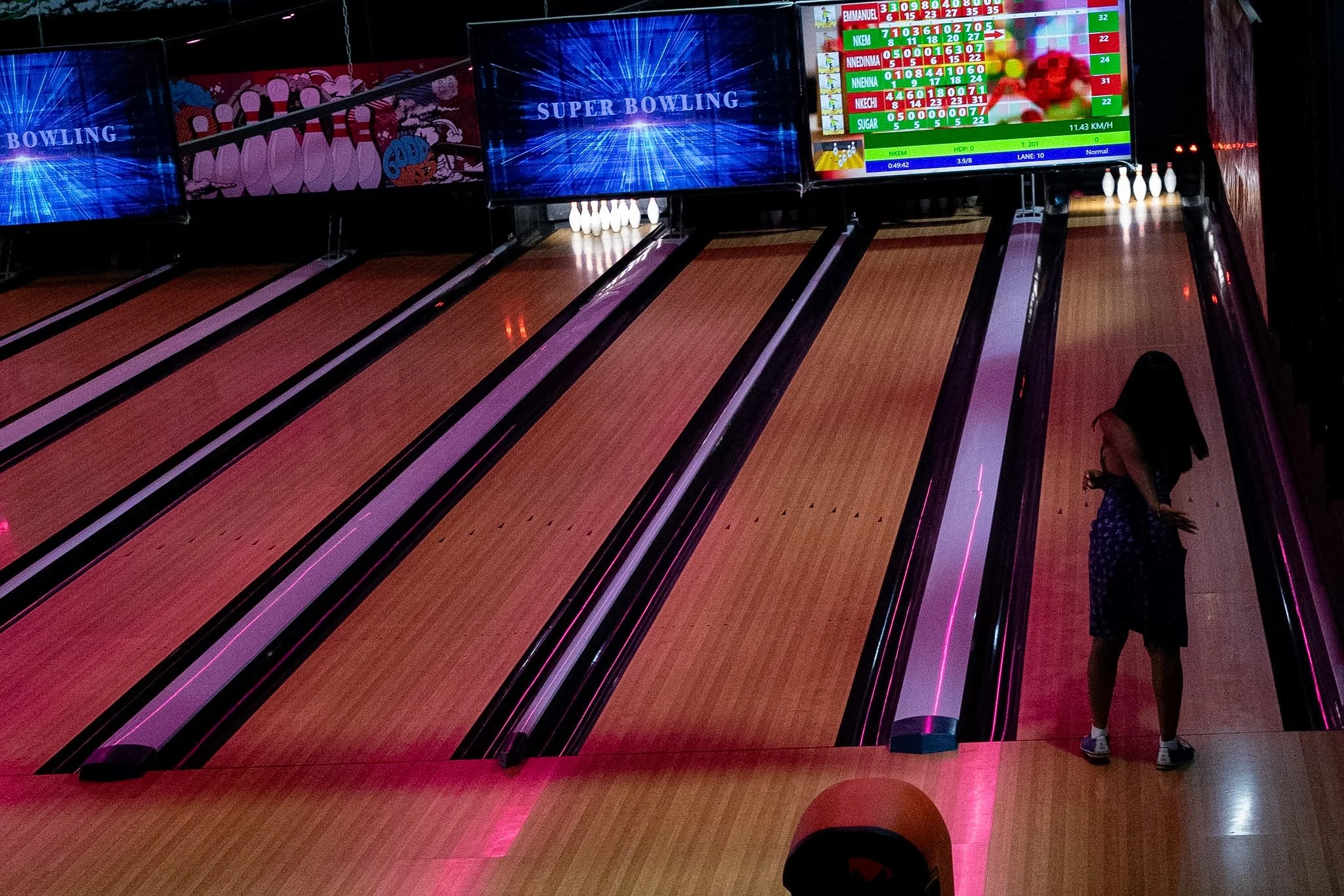 A bowling alley with three lanes, illuminated with pink and purple lights, and a woman standing near the right side of the lanes.