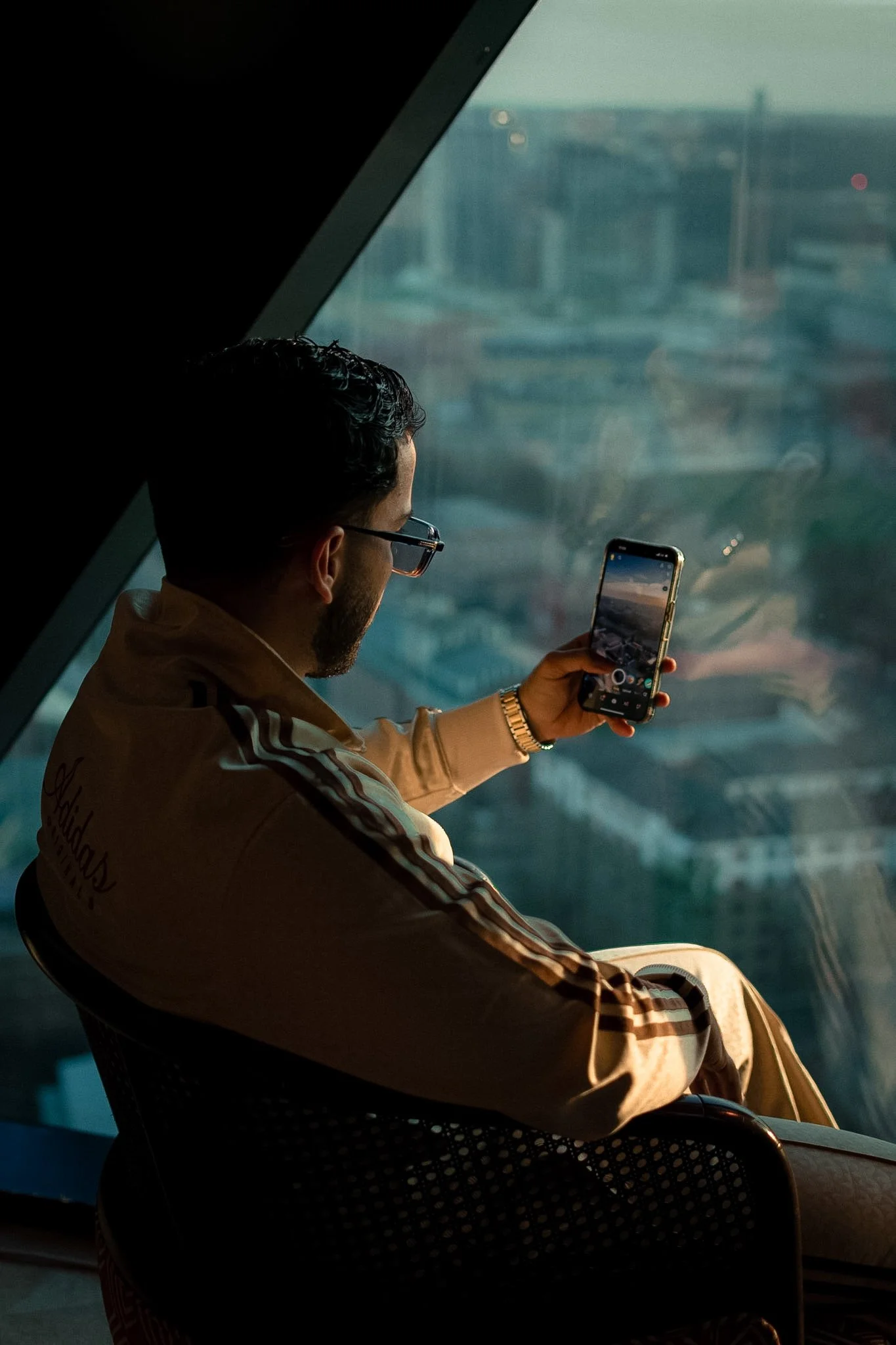 A man with dark hair and glasses wearing a beige jacket with striped sleeves, sitting on a chair and looking at his smartphone while inside a building with a large window showing a cityscape background.